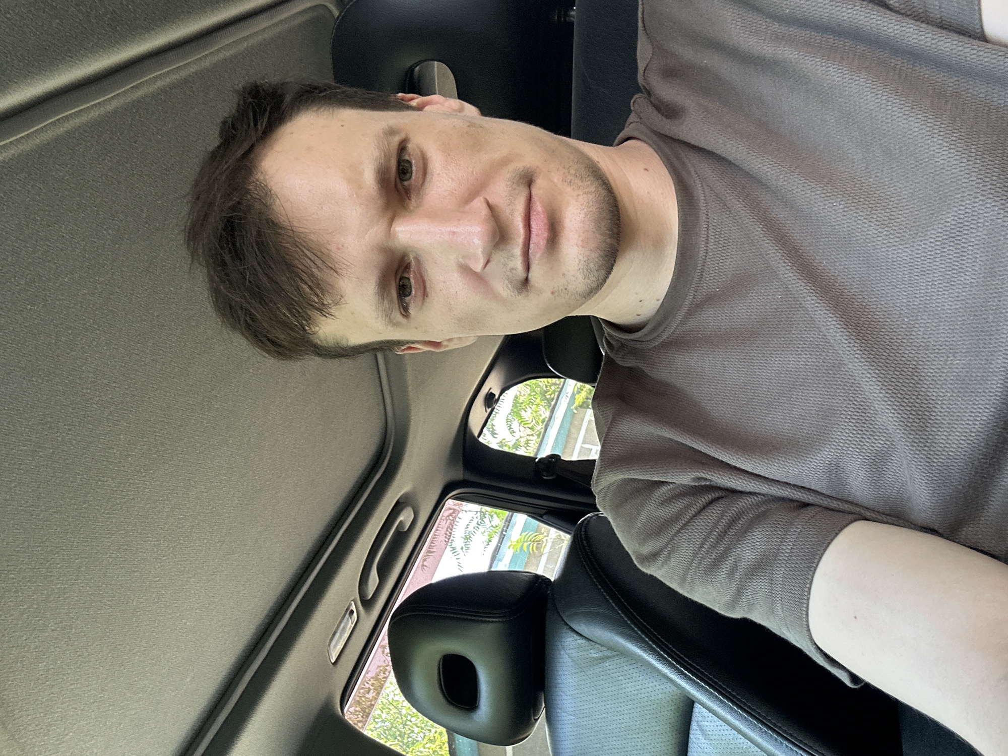 A medium shot captures a man sitting in the passenger seat of a car, looking directly at the camera. The interior of the car is visible, with a dark headrest of a seat to the left and the headrest of the driver's seat behind him. The car's ceiling is also visible, with a sunroof. Outside the car windows, trees and foliage can be seen, suggesting an outdoor setting, possibly in Makiv, Ukraine. The man has short, brown hair and is wearing a brown t-shirt. His expression is neutral. The lighting suggests it is daytime.