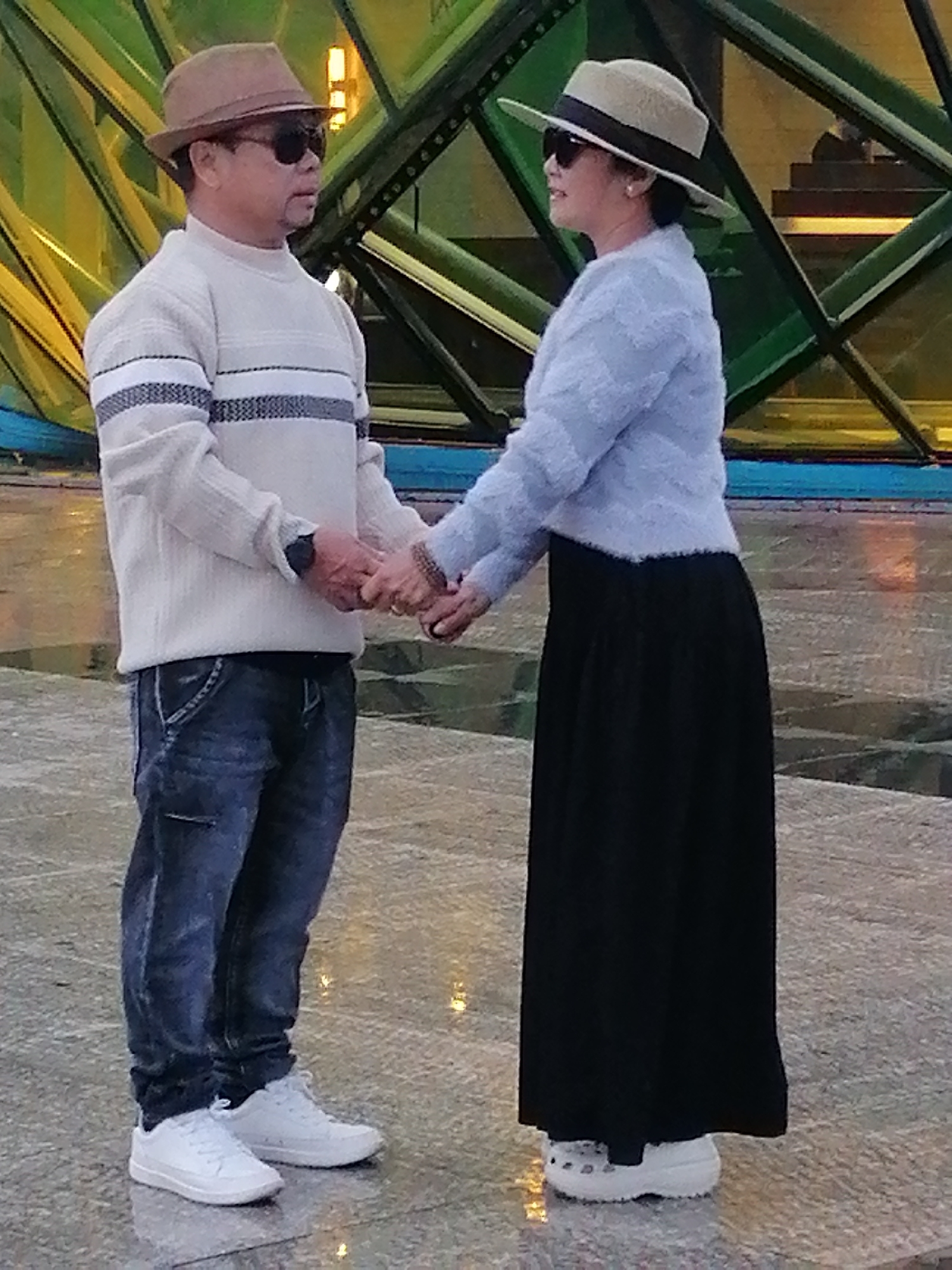 A couple stands holding hands in front of a modern structure with a green lattice. The man on the left wears a tan fedora, sunglasses, a cream-colored sweater with horizontal stripes, dark wash jeans, and white sneakers. The woman on the right wears a wide-brimmed hat, sunglasses, a light blue fluffy sweater, and a long black skirt with white clogs. The ground is paved with reflective tiles, and the lighting suggests it is either dusk or evening. The background structure appears to be an outdoor art installation or architectural feature. The location is identified as the Sculpture Garden of the Dalat Flower Park, Da Lat, Vietnam.