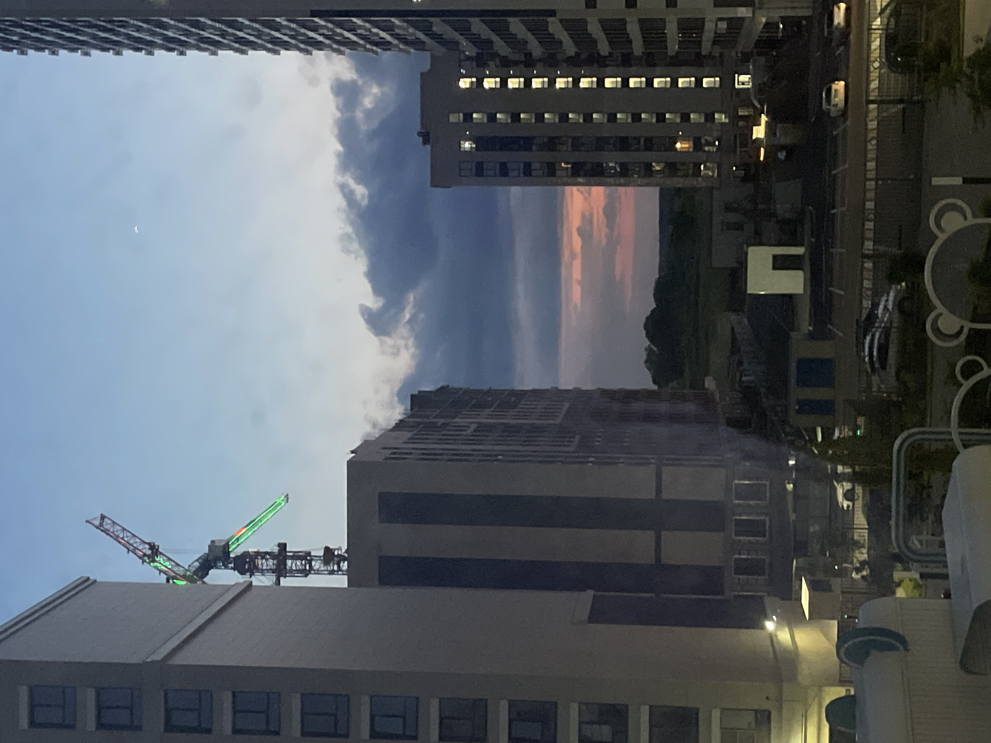 An exterior, eye-level, medium shot captures a construction scene at dusk in Stavropol, Russia. The foreground is dominated by the left side of a tall, beige apartment building with multiple stories visible, each featuring rows of dark-framed windows. To the right of this building, a large construction crane with green and red lights is prominently displayed against the twilight sky. Beyond these buildings, a series of apartment blocks with illuminated windows can be seen, suggesting an active residential area. The sky exhibits hues of blue, gray, and pink, indicative of sunset, with dark clouds gathering. In the distance, a treeline and a low horizon are visible. The ground level shows a parking lot with several parked cars and a road, hinting at the urban environment.