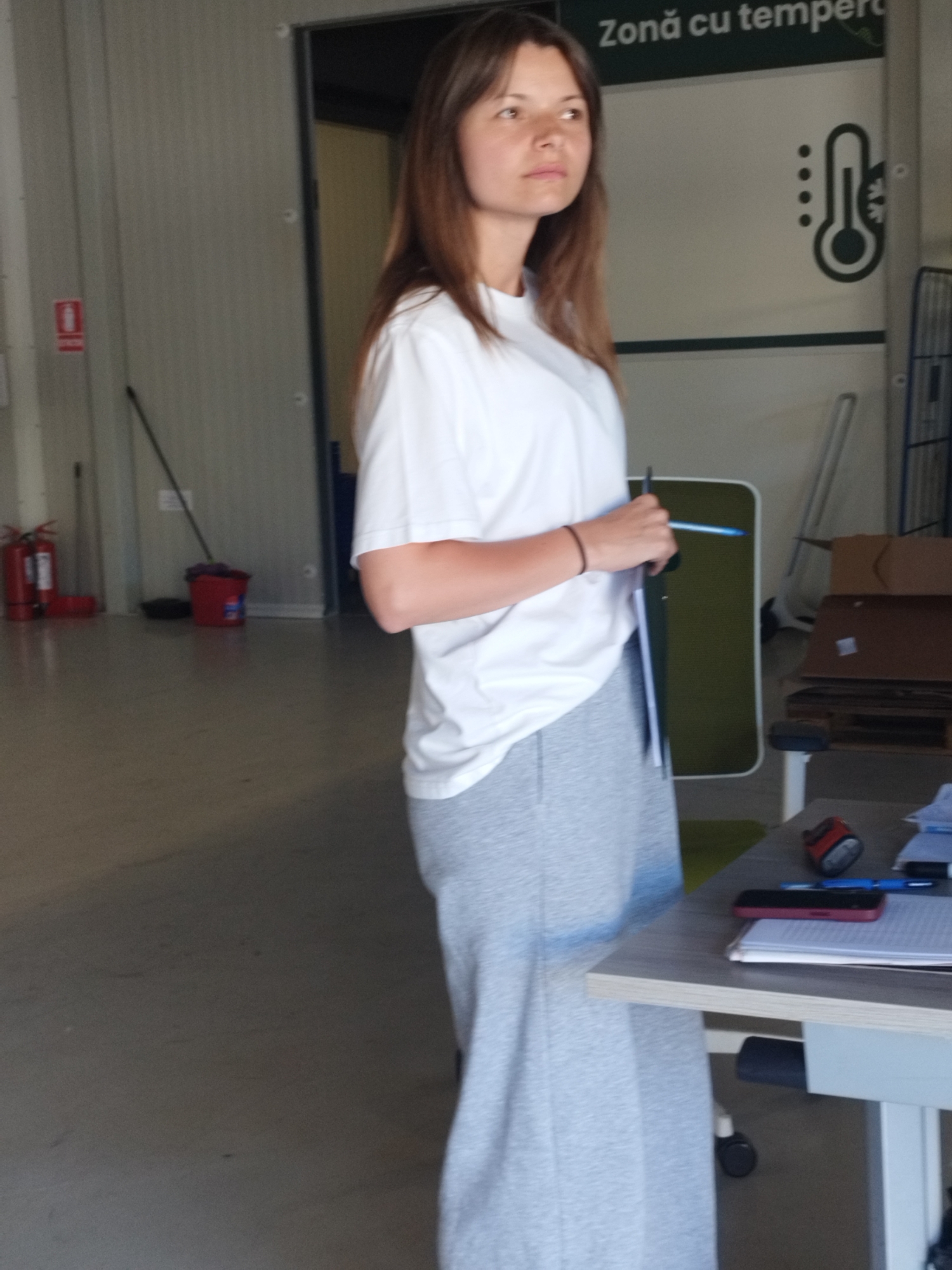 A young woman with shoulder-length brown hair stands in a room with concrete floors and grey metal walls. She is wearing a white t-shirt and grey sweatpants. Her left arm is bent and she holds a blue pen and a clipboard. Behind her, on the wall, is a sign with a thermometer icon and text in Romanian that translates to "Temperature controlled zone." To her left, two red fire extinguishers are mounted on the wall, and a red bucket with a mop is on the floor. To her right, a table with a laptop, papers, a red object that appears to be a stapler, and a smartphone is visible. The overall environment appears to be a storage or industrial area. The image was taken in Cernica, Romania.