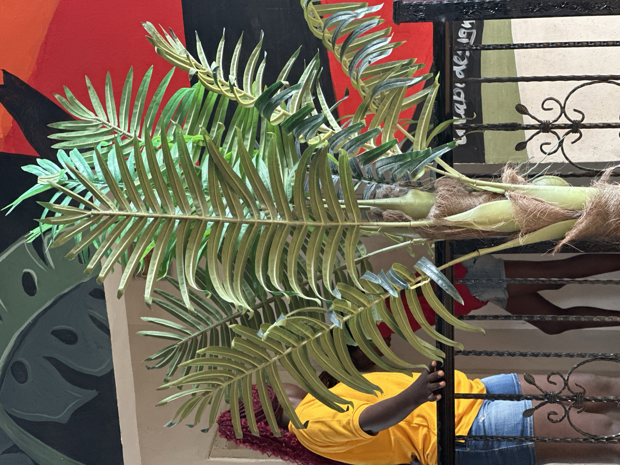 In Howayi, Nigeria, a close-up shot captures a section of a balcony with a large artificial palm plant prominently displayed. Behind the plant, two individuals are partially visible. One person, wearing a yellow t-shirt and denim shorts, stands on the left, leaning on the railing. Their dark hair is styled in vibrant red braids. To the right and further back, another person is seen wearing a red t-shirt and denim shorts, also standing near the railing. The background features a colorful mural with large green leaf shapes on the upper left and a striking red and black geometric pattern on the right. The railing is made of black wrought iron with ornate scrollwork. The scene appears to be outdoors, possibly a hotel or residential balcony, given the presence of a railing and the decorative mural.