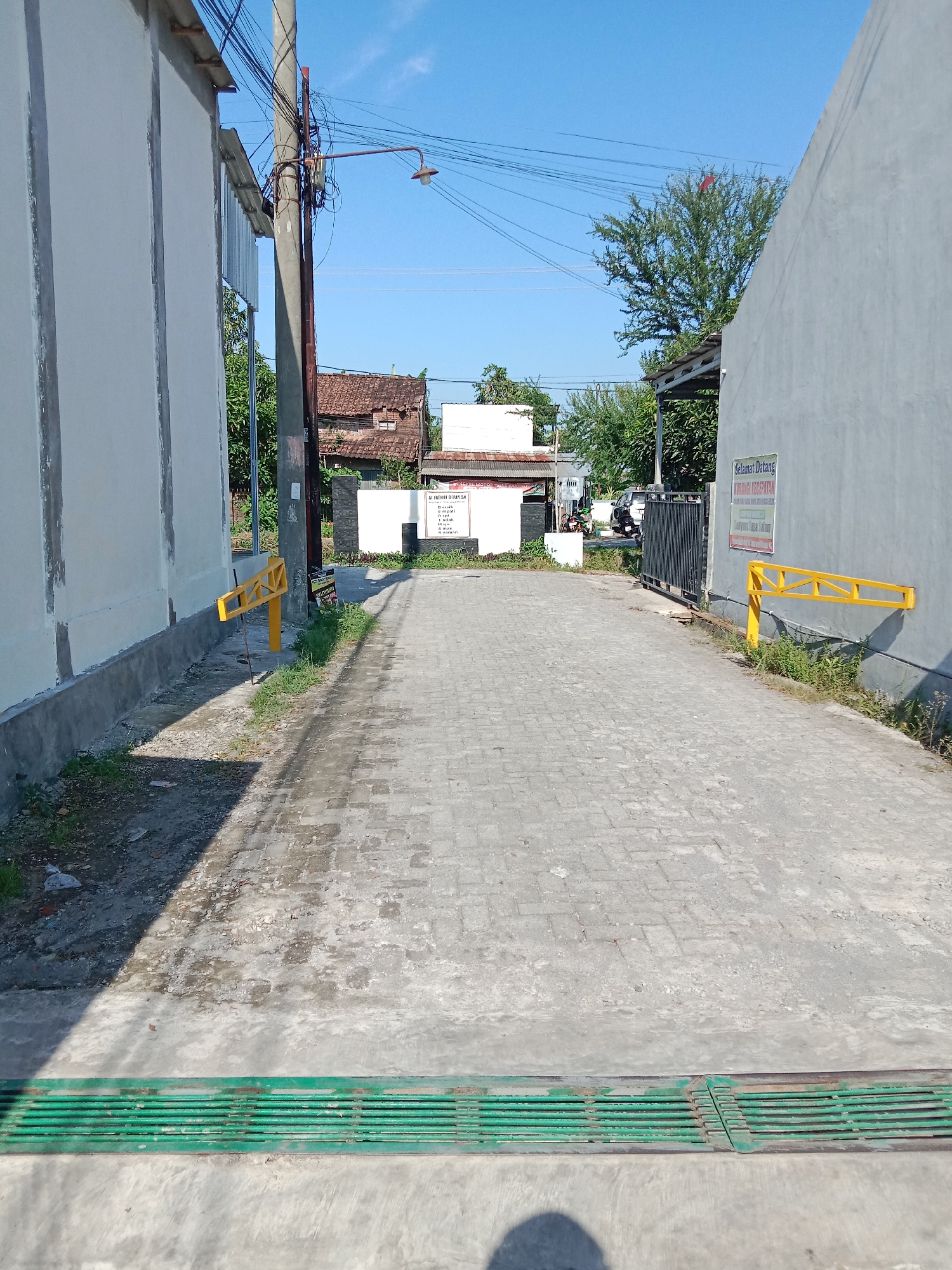 The image depicts a paved road located in Brangsong, Indonesia. The road is flanked by gray walls on either side with yellow boom barriers at both entrances. A green metal drain is visible in the foreground. Overhead power lines are visible. In the background, there are buildings with brown tile roofs, interspersed with trees and shrubs. The sky is clear and blue. The road surface appears to be constructed of interlocking pavers.
