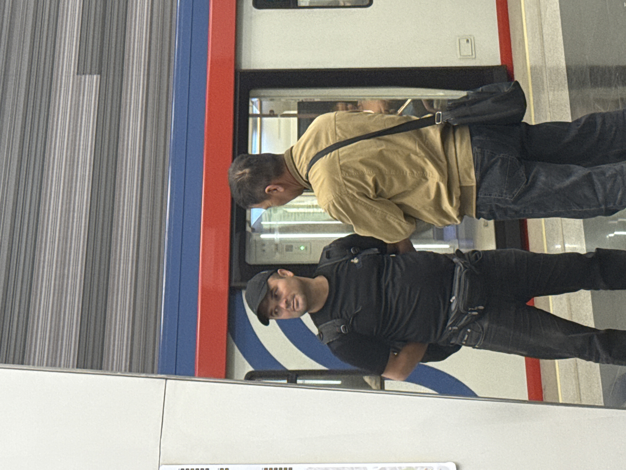 Two men stand near a train. One man wears a tan jacket and the other wears a black shirt and hat. The train has white, blue, and red stripes. The scene takes place in Moscow, Russia.