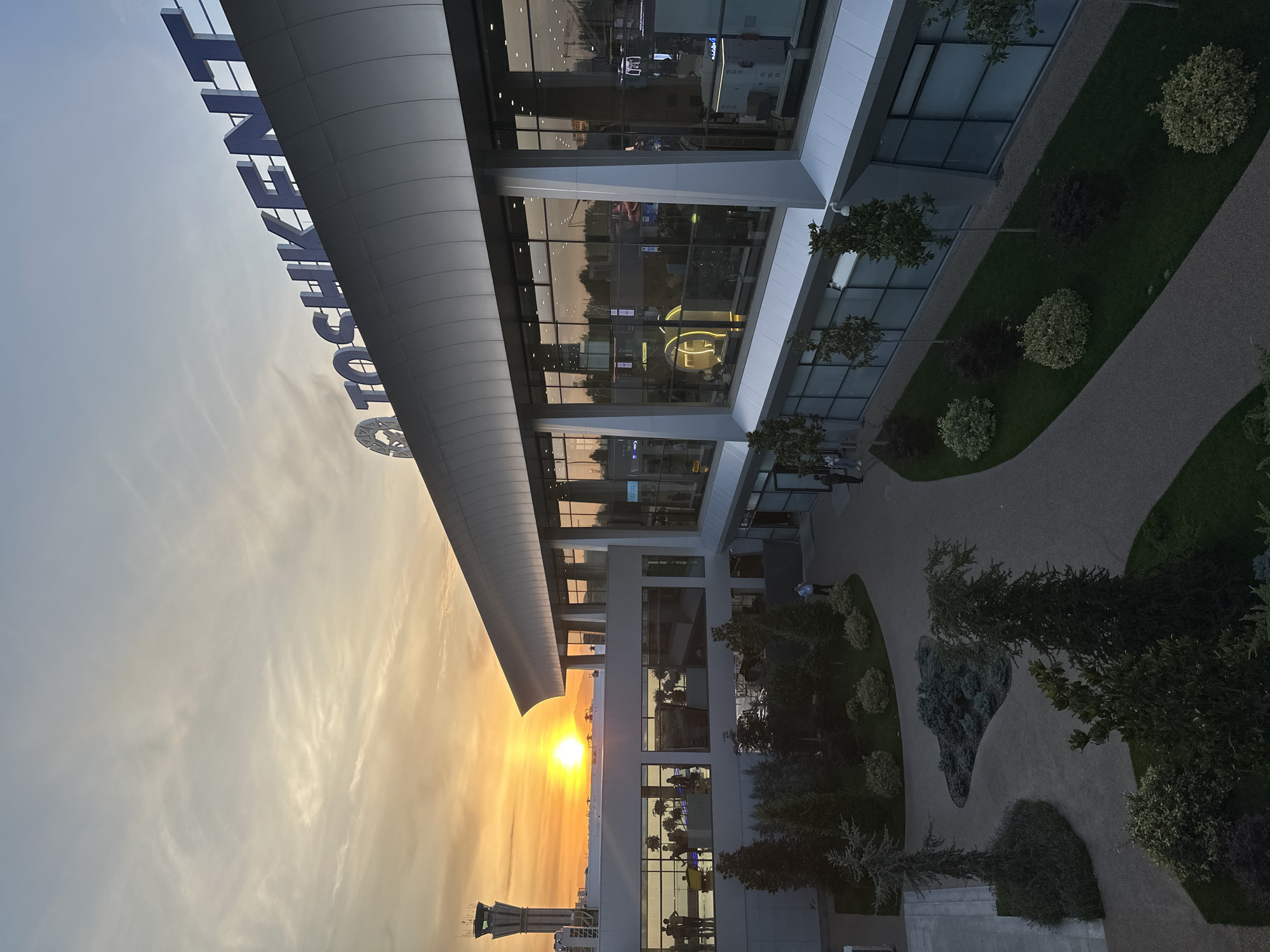 The image is a view of the Tashkent City district in Tashkent, Uzbekistan, at sunset. The sun is visible in the sky, shining brightly, and its light is reflected in the windows of a modern building. The name "Tashkent City" is seen in large letters on top of the building. The building itself has a sleek, modern design with a curved roofline. A control tower-like structure is visible in the distance. There are people inside the building, with their silhouettes visible in the windows. Outside the building, there is a landscaped area with trees, shrubs, and a walkway.