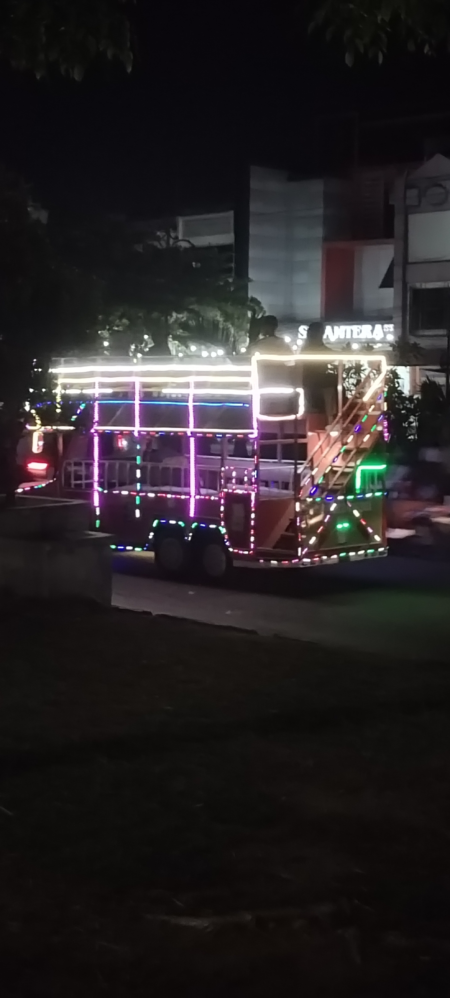 At night in Lhokseumawe, Indonesia, an elaborately decorated truck adorned with colorful lights drives down a road. The truck's frame is outlined with strings of white, blue, pink, and green lights, creating a festive appearance. Two people stand on the top level of the truck. A building with illuminated signage is visible in the background. The foreground is dark and shows a grassy area.
