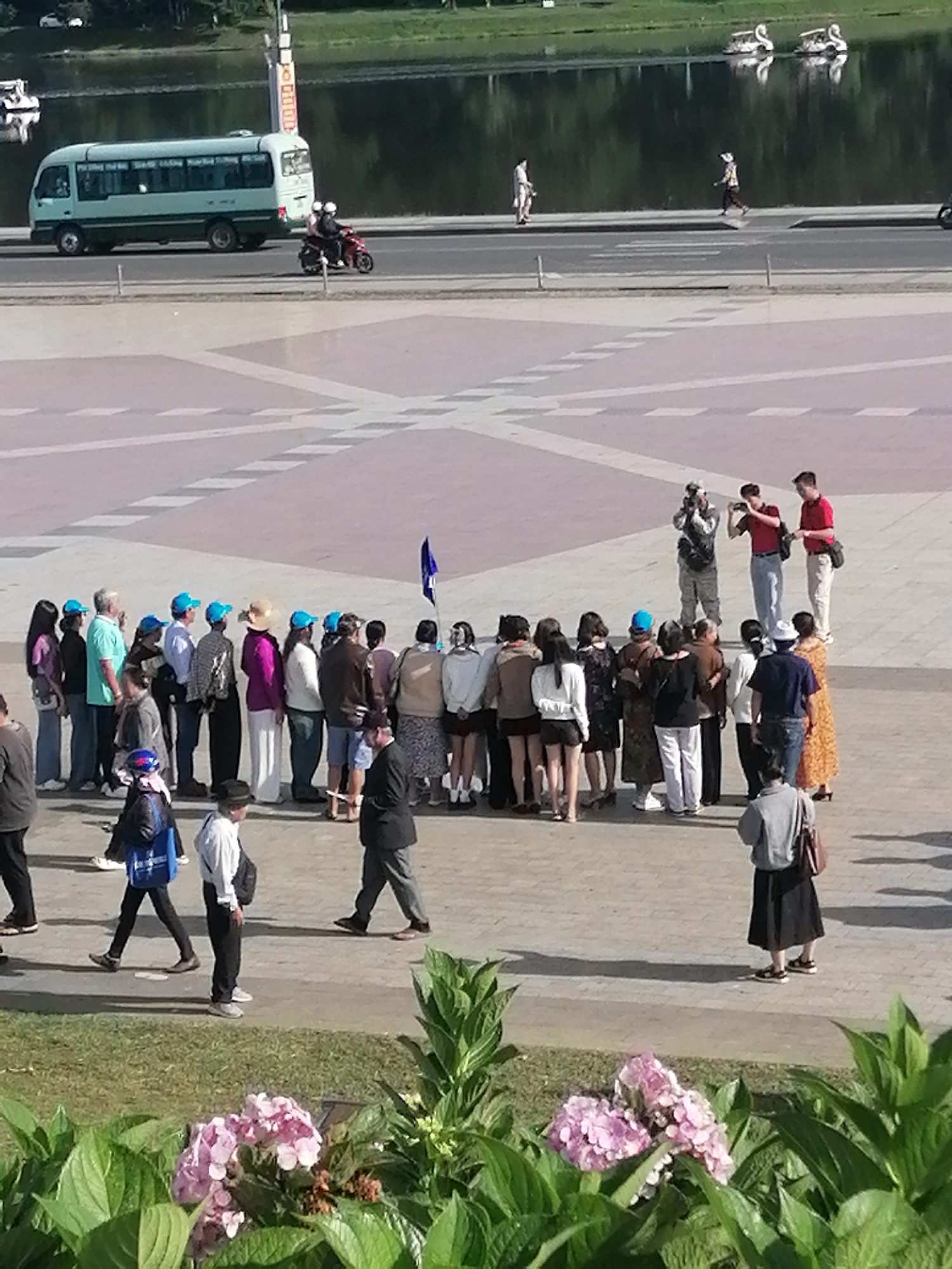 This image depicts a scene near Xuan Huong Lake in Da Lat, Vietnam. In the background, a light blue bus is parked on the side of a road, with a motorcycle carrying two passengers also visible. The lake itself is calm, reflecting the green of the surrounding trees. On the paved area in the foreground, a group of people are gathered. Some are wearing blue hats, while others are dressed in casual attire. Three individuals appear to be taking photographs. The group is diverse, with men and women of various ages. The ground is paved with light-colored tiles, and shadows from the people and objects are cast across the surface, indicating a sunny day. In the bottom right of the frame, a cluster of pink hydrangeas is in bloom, adding a touch of natural beauty to the urban setting.