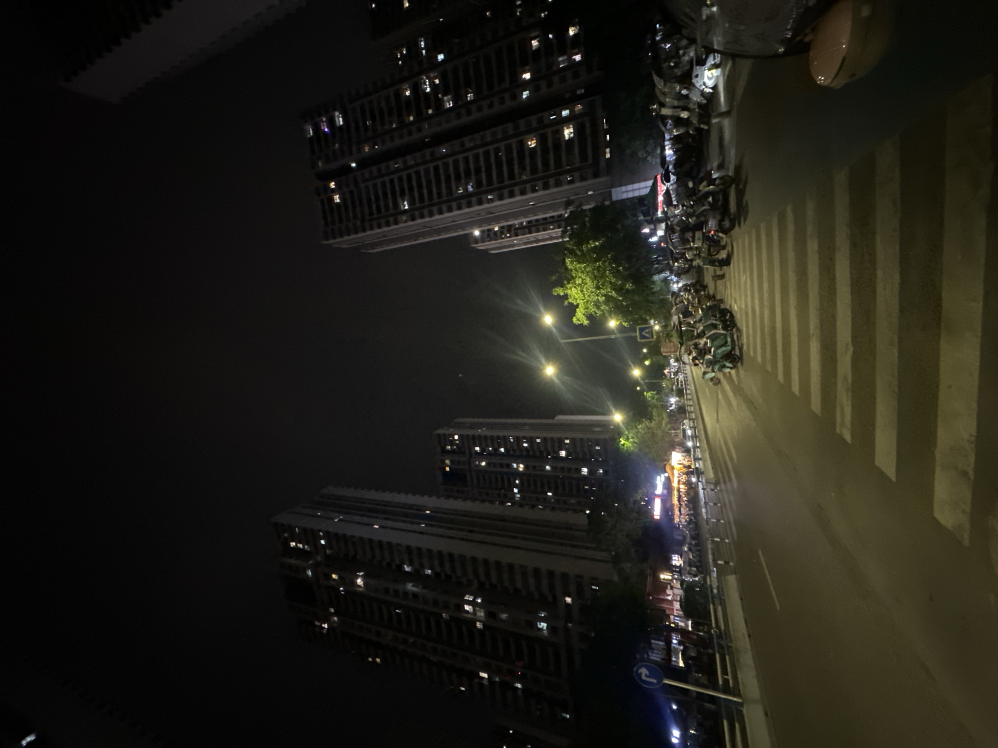 The image depicts a nighttime street scene in the Chongqing High-tech Zone, China. Tall apartment buildings stand on either side of the street, illuminated by interior lights. Streetlights line the roadway, casting a warm glow. Numerous scooters are parked along the curb. A crosswalk stretches across the road. Overall, the scene conveys a sense of urban life at night.