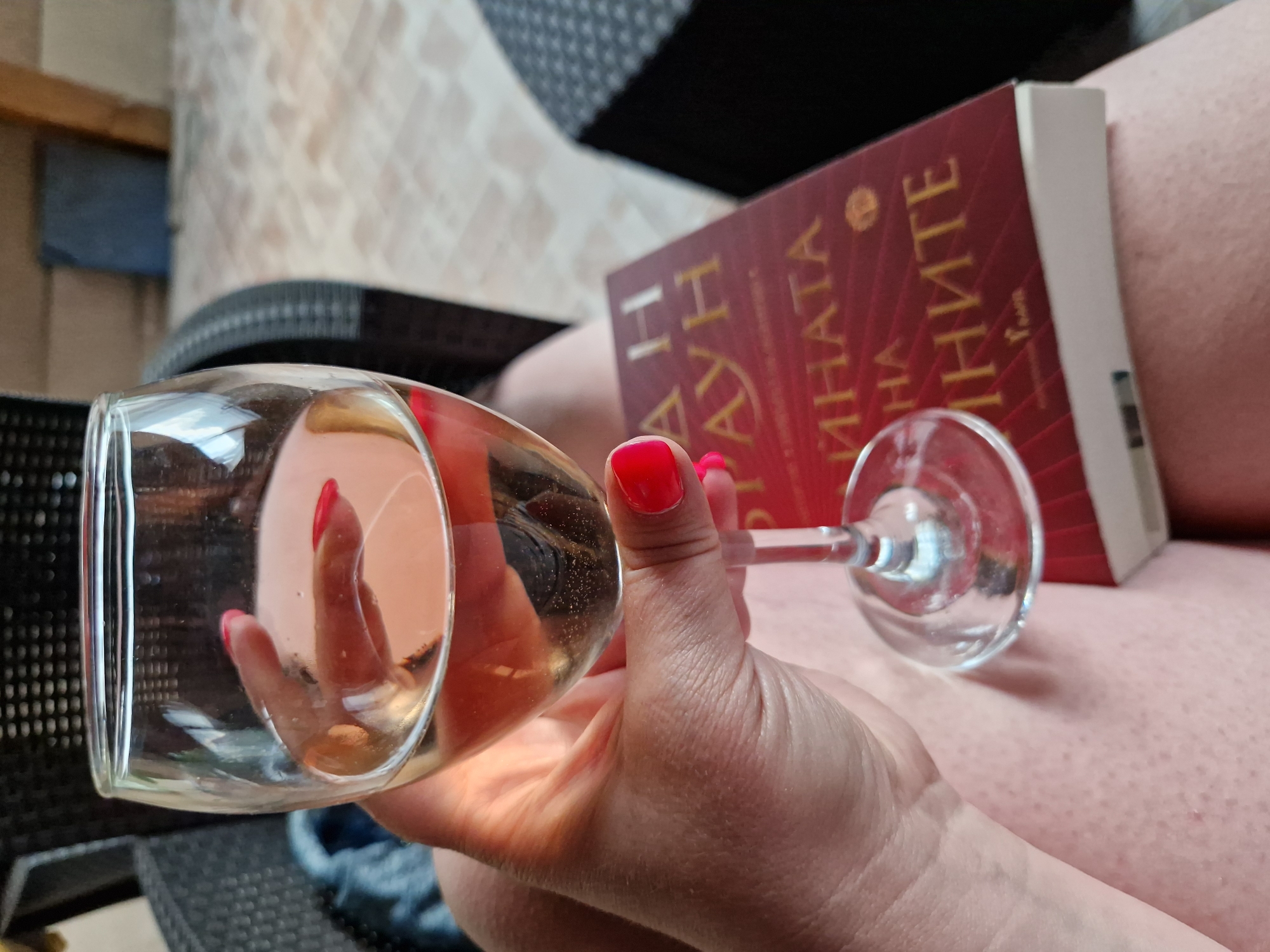 A person is holding a wine glass filled with a light-colored beverage. The person's hand, with red nail polish, is visible supporting the glass. In the background, a book with a red cover and gold lettering is resting on the person's lap. The environment appears to be outdoors, possibly a patio or garden area, with textured flooring and outdoor furniture. The lighting suggests it is daytime. The specific location in Sofia, Bulgaria, cannot be determined from the visual information provided.