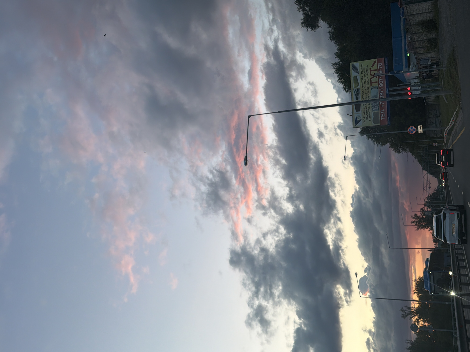 The image depicts a street scene in Samara, Russia, at dusk. A highway stretches into the distance, with multiple lanes of traffic visible. Several cars and a bus are present on the road. Streetlights illuminate the scene. On the right side of the road, a billboard advertises various construction materials, including pipes, metal, and building panels. A phone number is clearly visible on the billboard. A small building, possibly a service station or shop, is located near the billboard. A few pedestrians are standing near the building. The sky above is filled with dramatic clouds, tinged with pink and orange hues, indicating the setting sun. Trees line the right side of the road, and foliage is visible on the left.