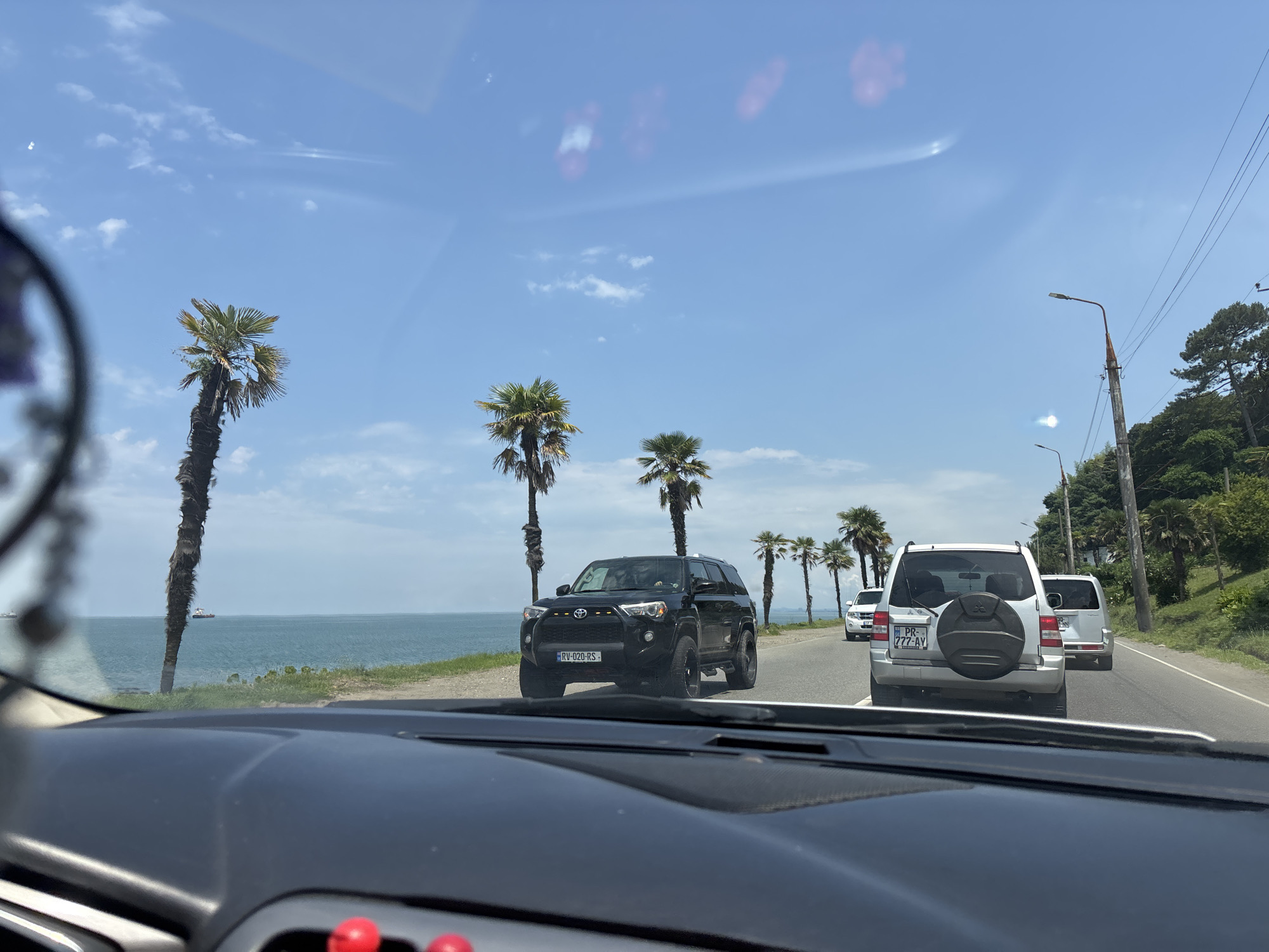 The image captures a view from inside a vehicle driving along a coastal road in Batumi, Georgia. The foreground shows the interior of the car, including a dashboard with red accents. The road ahead is lined with palm trees and leads towards the Black Sea, which is visible on the left side of the frame. Several vehicles are on the road, including a black SUV with a license plate reading "RV-020-RS" and a silver SUV with a spare tire mounted on the back and a license plate that appears to read "PR-777-AV". In the distance, ships can be seen on the water. The sky is blue with scattered clouds, and sunlight creates reflections on the car's windshield. The environment appears tropical or subtropical, characteristic of Batumi's climate.