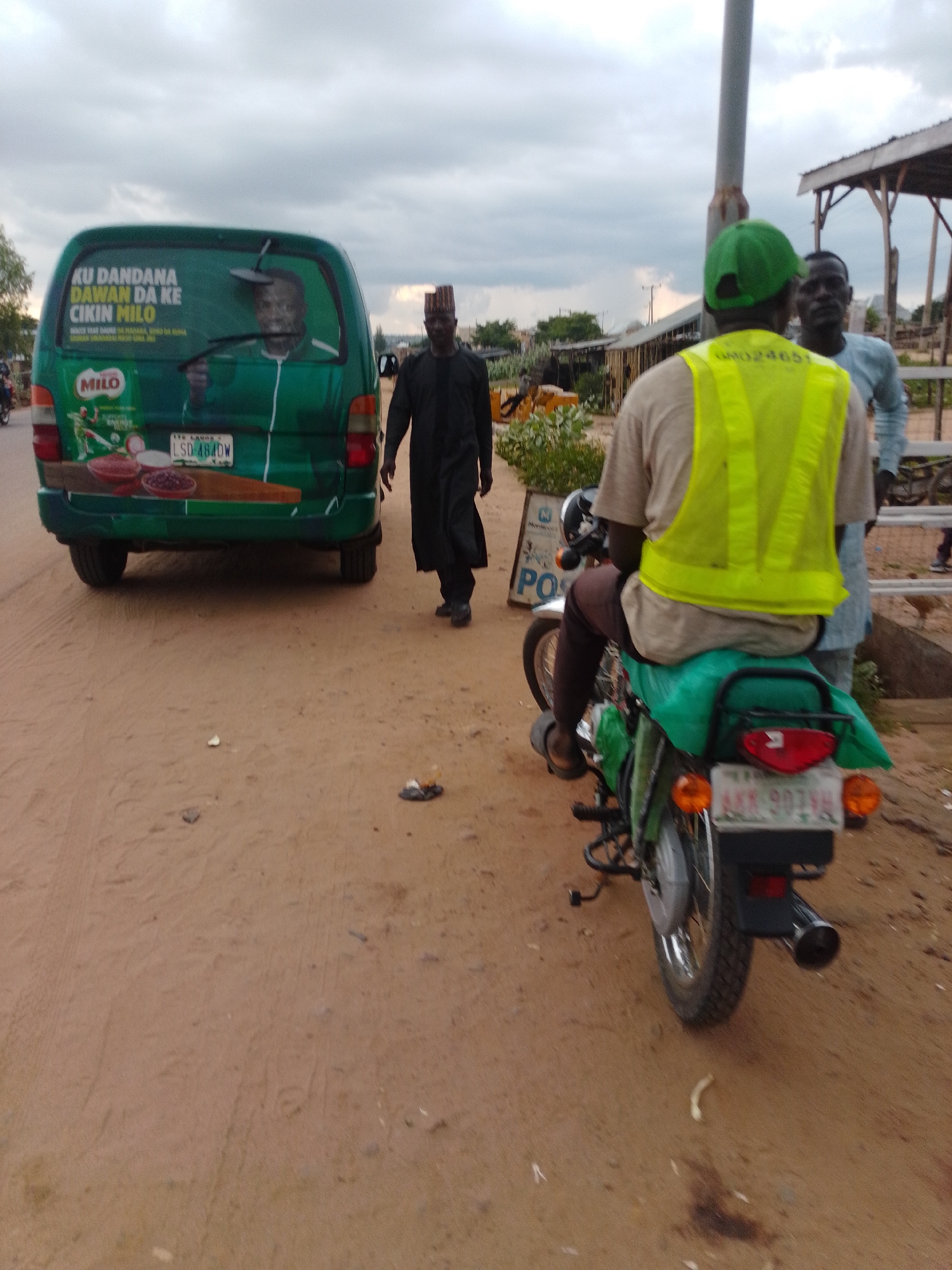 The image, taken on a dirt road in Gombe, Nigeria, depicts a street scene with a green van adorned with political campaign posters, a man in traditional attire walking away from the van, and a person on a motorcycle wearing a high-visibility vest. The van has an advertisement for Milo, a popular beverage. The motorcycle, parked to the right, has a license plate that reads "AKR 907VH." The background shows a cloudy sky and the edge of a structure, possibly a market or roadside stall. The ground is covered with reddish-brown dirt.