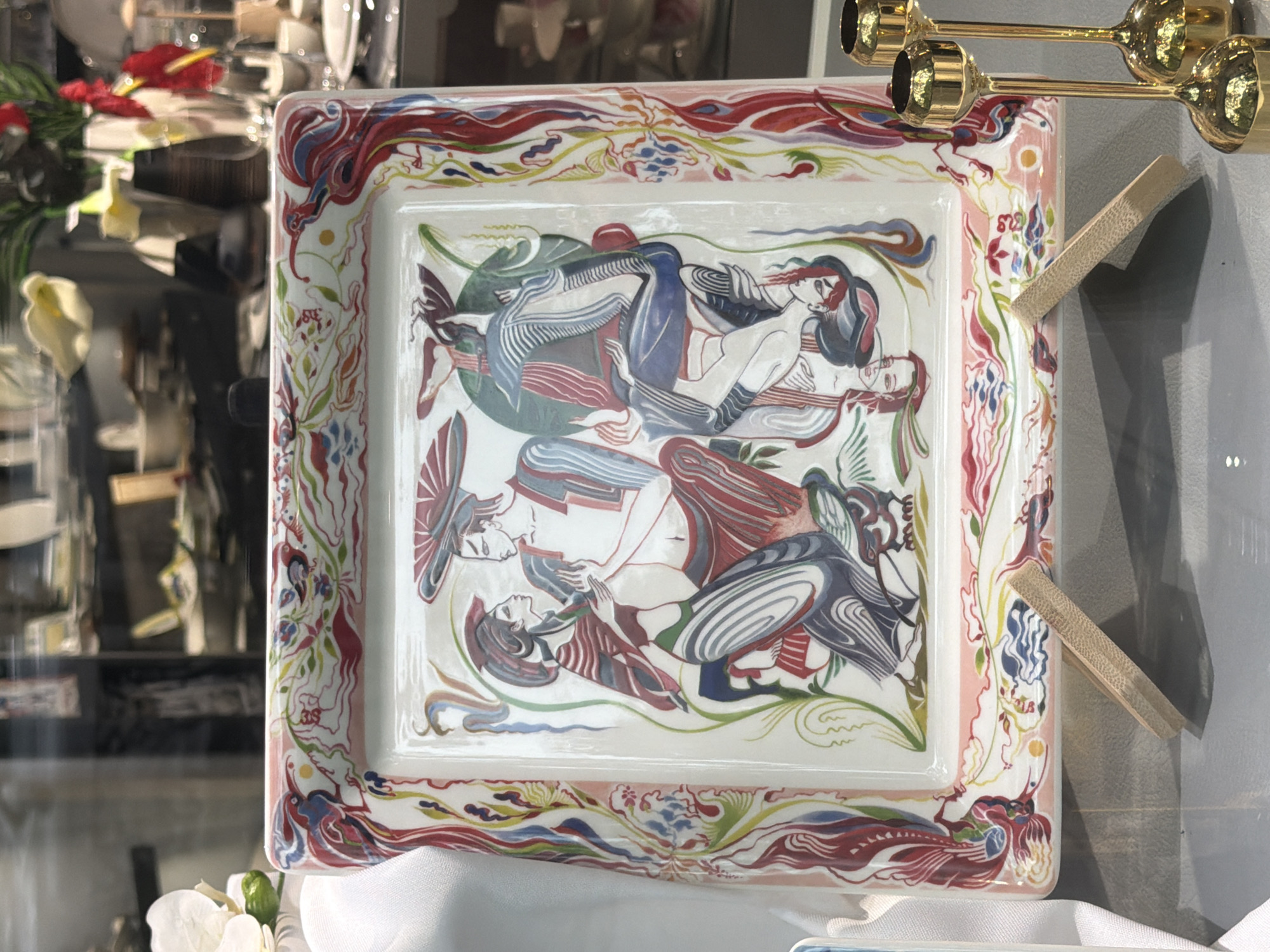 The image shows a square ceramic plate with an ornate, colorful border featuring stylized floral and avian motifs in red, blue, green, and yellow. The central image on the plate depicts three figures rendered in a flat, stylized artistic manner, with bold outlines and block colors. Two figures are positioned centrally, facing each other, with one figure partially embracing the other. A third figure appears to be positioned upside down or in a contorted pose below the central figures. The figures are dressed in clothing with geometric patterns, and one prominent detail is the large, conical hats worn by two of the figures. The plate is displayed on a light-colored stand made of wooden slats, positioned in what appears to be a retail display. In the background, shelves filled with various ceramic and decorative items are visible, suggesting a shop or gallery setting. Further in the background, out-of-focus elements indicate a broader environment, possibly with other displays and lighting. The overall impression is of an art piece or decorative item being showcased.