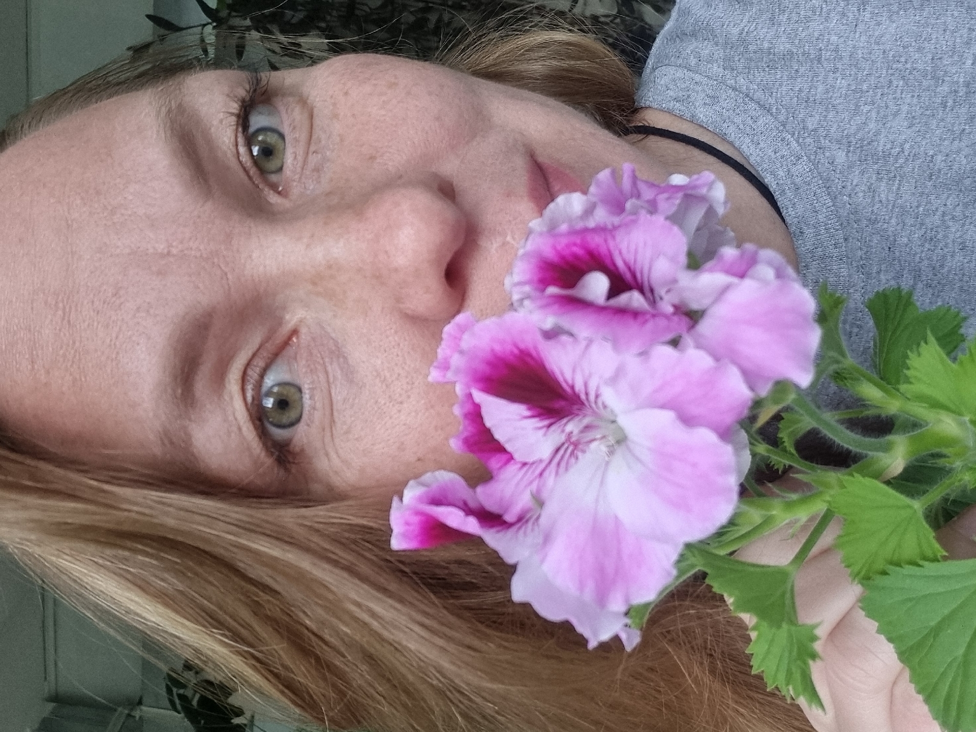 A close-up, slightly angled portrait shows a fair-skinned individual with freckles and light-colored hair. They have green eyes and are positioned with their face close to the camera. A bouquet of pink and purple geraniums is held near their face, partially obscuring their mouth. The person is wearing a gray t-shirt with a black cord around their neck. The background is indistinct, with some green foliage visible at the top left corner. There is no evidence to suggest the location is in the city of Kemul, Russia.