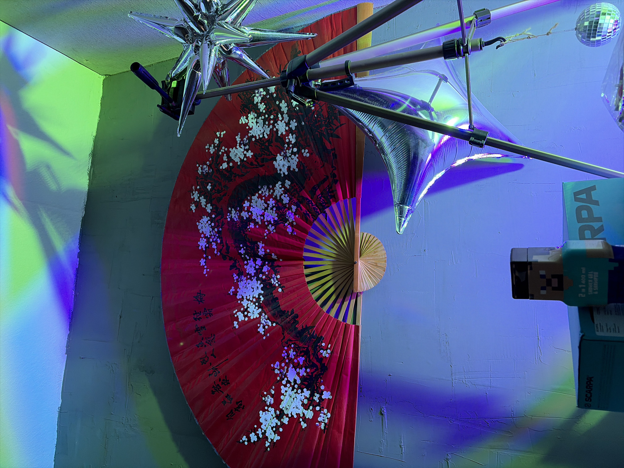 The image shows an indoor scene illuminated by colored lights, casting shadows and reflections. In the foreground, a red fan with a cherry blossom motif is displayed. To the right of the fan is a tripod with various objects attached, including a metallic star-shaped decoration and a silvery, balloon-like object. Below these items, two boxes are visible, one labeled "SCARPA" and another with text and a pixelated character. A small disco ball hangs from a string on the tripod. The background consists of a textured wall illuminated with blue and green lights, with abstract light patterns projected onto it. The scene appears to be a decorative setup, possibly for an event or artistic display. There are no people or any clear indicators of the specific location, including the city of Moscow, Russia.