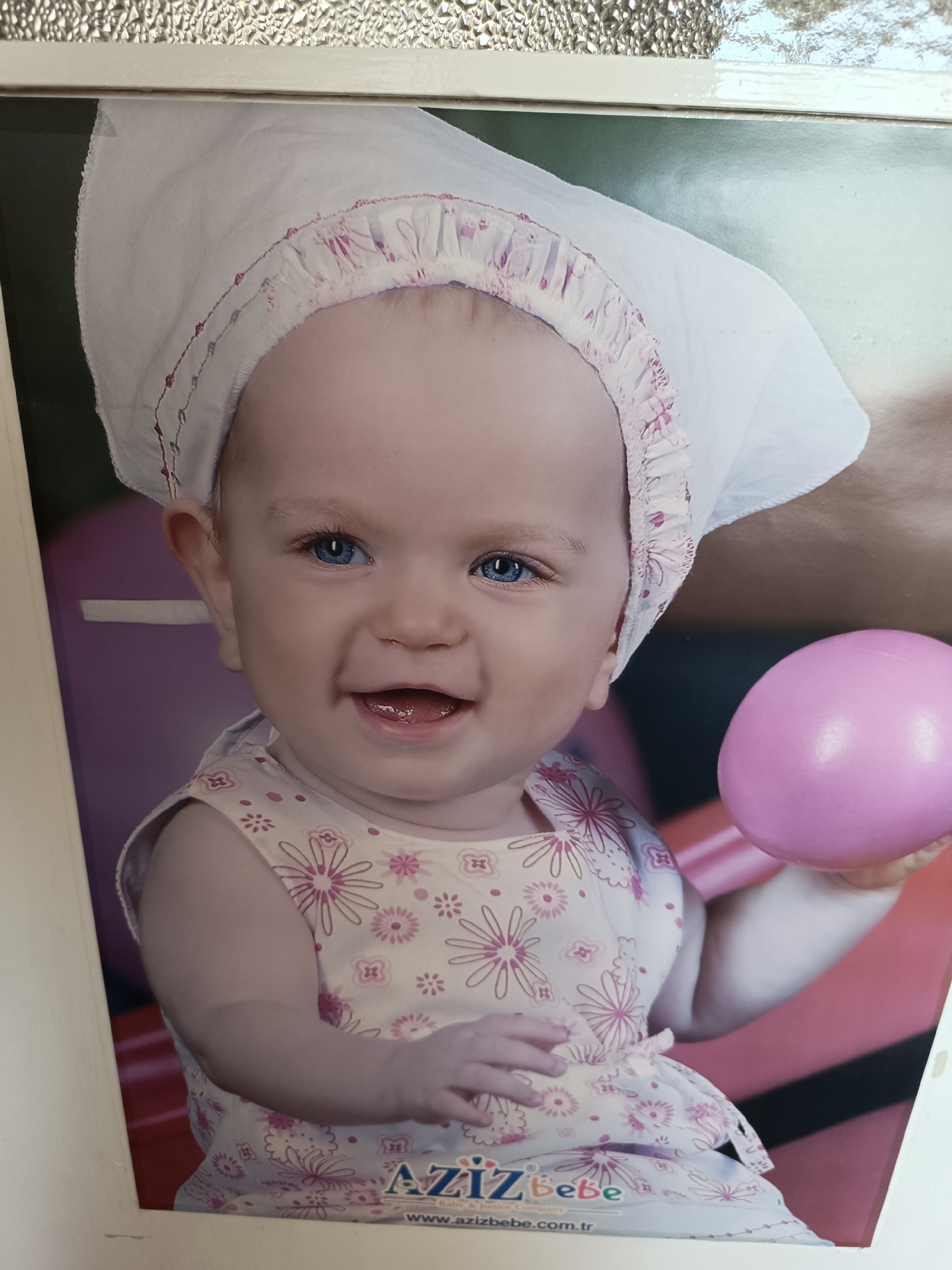 The image is a photograph of a poster featuring a baby. The baby has fair skin, blue eyes, and is wearing a white bonnet with a ruffled, pink floral trim and a dress with a similar floral pattern. The baby is smiling and holding a pink, egg-shaped object. The background features a purple and red striped surface. At the bottom of the poster, the text "AZIZ Bebe" is printed, along with the website address "www.azizbebe.com.tr". The location of the image is not provided and cannot be determined from the image.