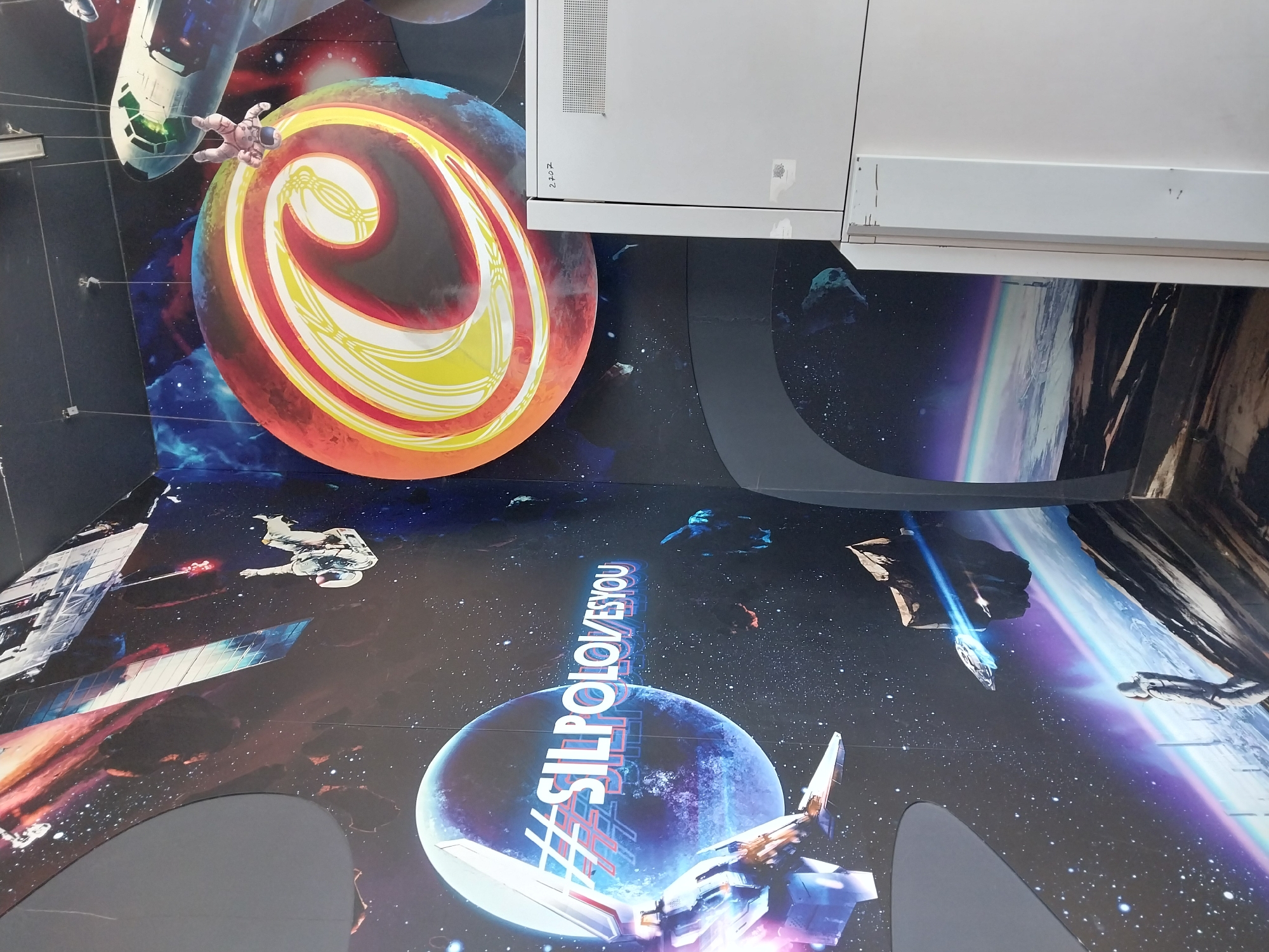 The image depicts a wall mural with a space theme. The mural features planets, stars, spaceships, and astronauts. A prominent feature is a large blue planet with the hashtag "#SILPOLOVESYOU" written across it in white lettering with a red outline. To the right of the planet is a large, colorful, circular graphic with a stylized "C" in yellow and white. In the upper right corner, a white appliance, possibly a refrigerator or copier, is visible with the number "2702" written on it. The overall scene suggests an interior space decorated with a space-themed mural. The location is specified as Dnipro, Ukraine.
