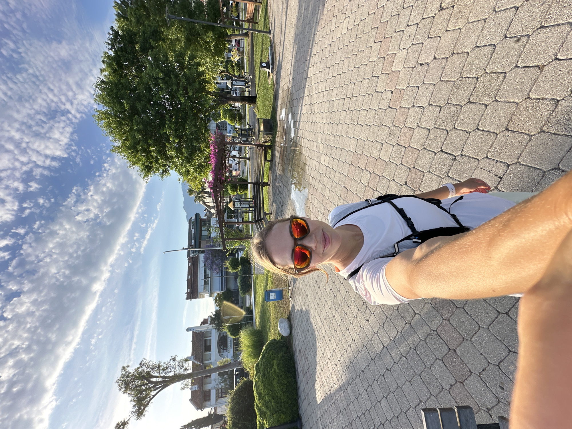 A selfie captures a woman in an outdoor setting, likely a park or promenade in Fethiye, Turkey, during late afternoon or early evening. The woman, with blonde hair, wears reflective orange sunglasses, a white t-shirt, and a black harness or straps. She holds the camera with her right arm, and a white band is visible on her left wrist. She presents a slight smile, suggesting a pleasant mood.
The environment features a light-colored, patterned block paved path, parts of which appear wet. Adjacent to the path are well-maintained green grass, manicured bushes, and trees casting long shadows. Several park benches are scattered throughout the green space. In the middle ground, traditional buildings with red-tiled roofs and light-colored facades are visible, and in the distance, large mountains rise under a bright blue sky. The sky is adorned with scattered white, wispy clouds. A large green tree and another with purplish-pink flowers near a wooden pergola are prominent natural elements. The scene conveys a sunny and serene atmosphere. No discernible text is visible.