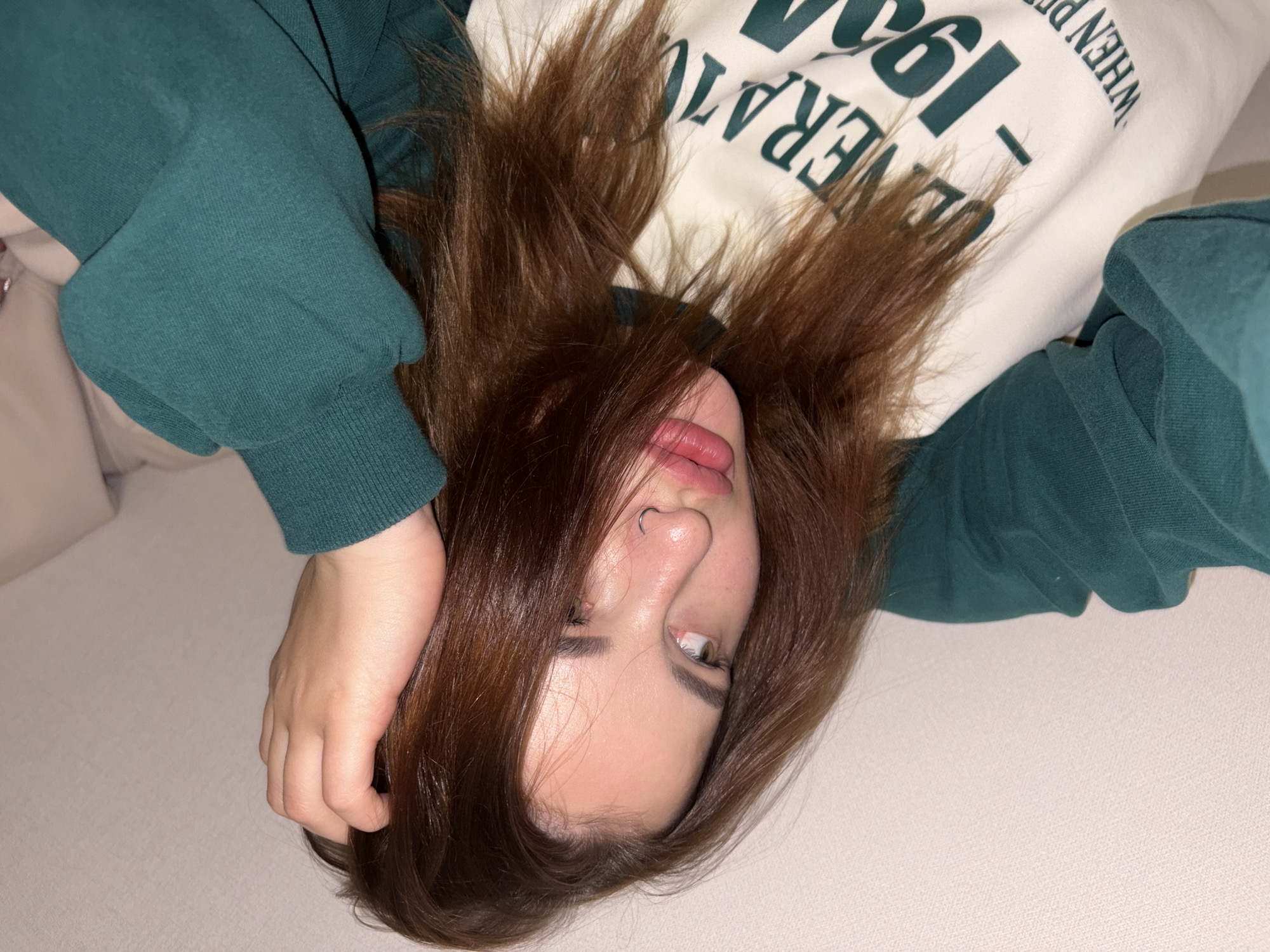 A close-up, rotated image features a young individual lying on a light, textured surface, likely a bed or couch, with their head positioned towards the bottom of the frame. The subject has long, reddish-brown hair that spreads around their head, partially covering their face. They are wearing a dark teal or forest green long-sleeved sweatshirt over a white top.
The person's face displays a playful or silly expression, with their tongue slightly extended and one eye (the left eye from the viewer's perspective) rolled upwards, revealing the sclera and a light-colored iris. A silver hoop nose ring is visible on their left nostril. Their right hand is casually resting on or near their hair, partially visible at the bottom left of the frame.
Visible text on the white under-top, in a dark green font, includes fragments such as "GENERATIO" and "WHEN P - 199". The background is minimalist, consisting of the light-colored surface, reinforcing the focus on the individual. The overall mood is casual and informal.
Location context: Unknown, Unknown
