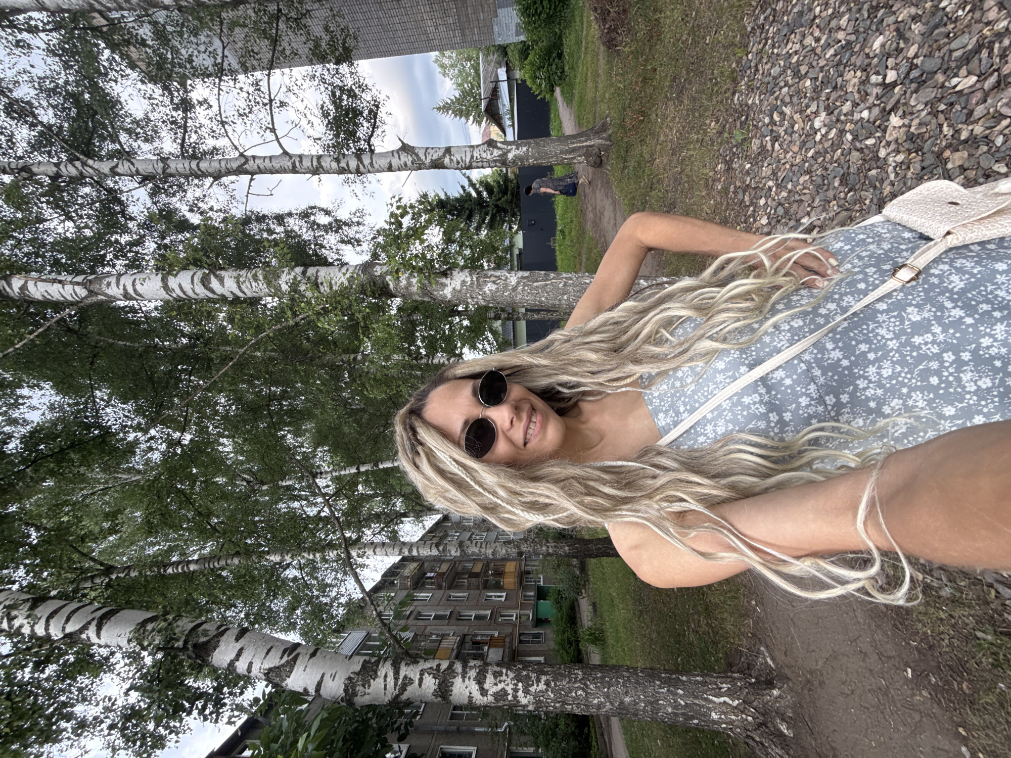 In this full-body selfie, a woman with long, blonde dreadlocks and sunglasses smiles at the camera. She is wearing a light blue dress with a white floral pattern and a straw crossbody bag. The background depicts an urban scene with birch trees, a grey apartment building, and a gravel path. In the distance, another woman walks away from the camera.