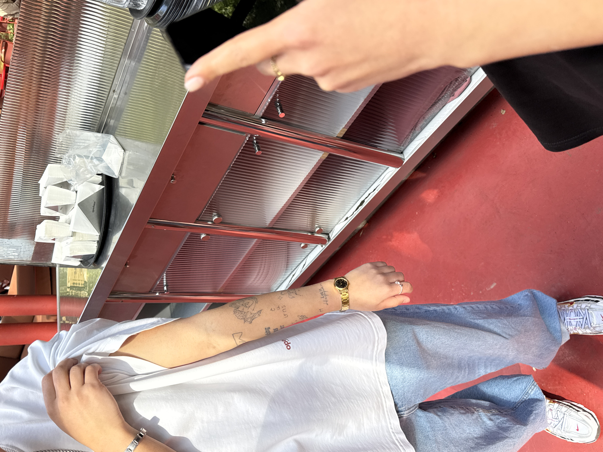A first-person perspective photograph shows a person wearing jeans, a white t-shirt, and silver and white sneakers standing on a red surface in Yerevan, Armenia. They have tattoos on their left arm and wear a gold watch and ring. Another person's hand, adorned with a ring, is visible in the upper part of the frame, holding a cell phone pointed towards a stainless steel structure, possibly a kiosk, with ribbed plastic panels. Packets or containers, some in clear plastic, are on the kiosk. The photograph is taken in daylight.