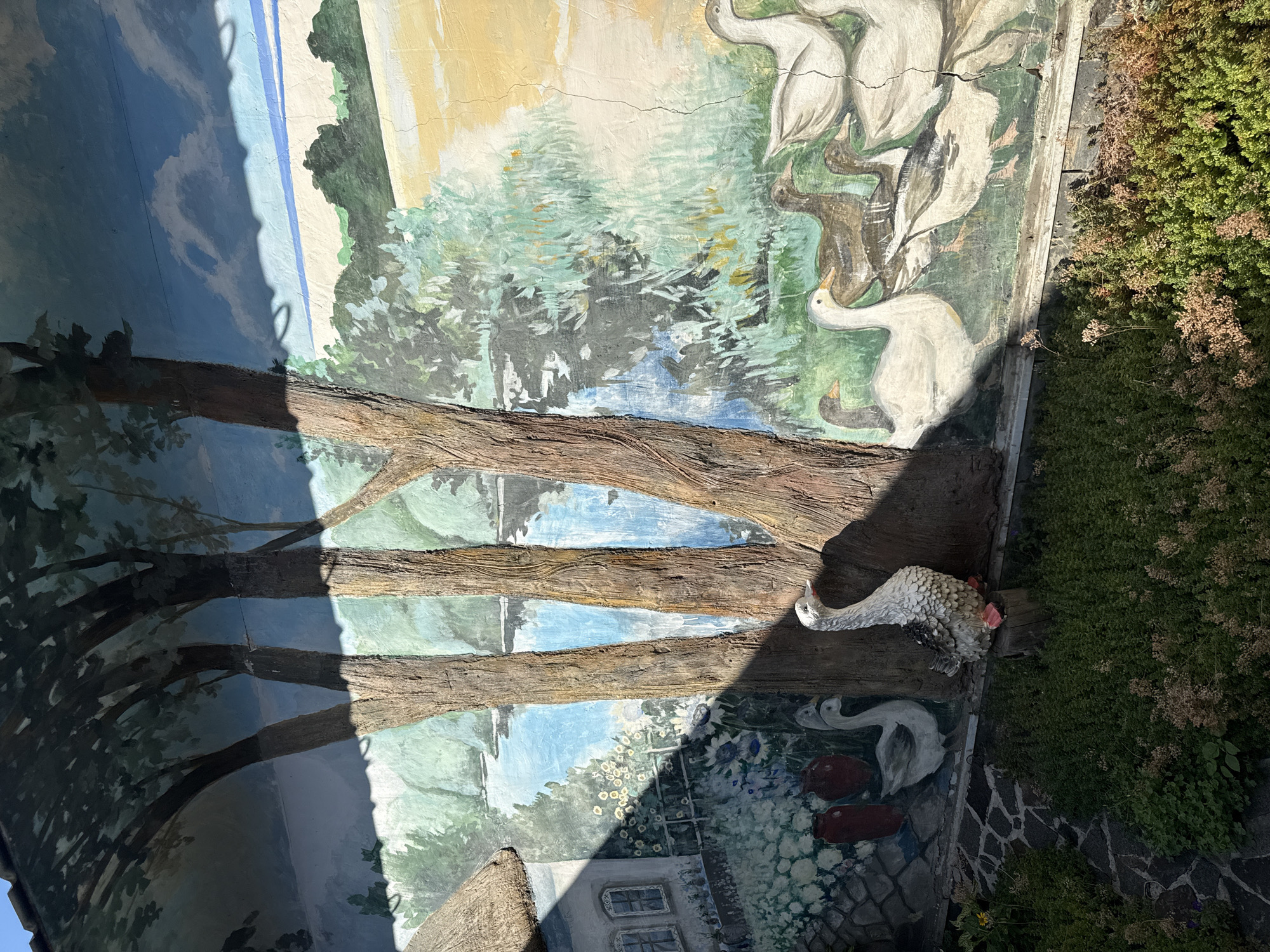 The image shows a mural on a wall, depicting a rural landscape with trees, a body of water, and several geese and ducks. In the foreground, there is a garden with green bushes and a decorative statue of a goose. The mural is partially covered by shadows from trees. The setting appears to be outdoors, with the mural painted on a textured wall. The provided context indicates this scene is located in the city of Okhtyrka, Ukraine.