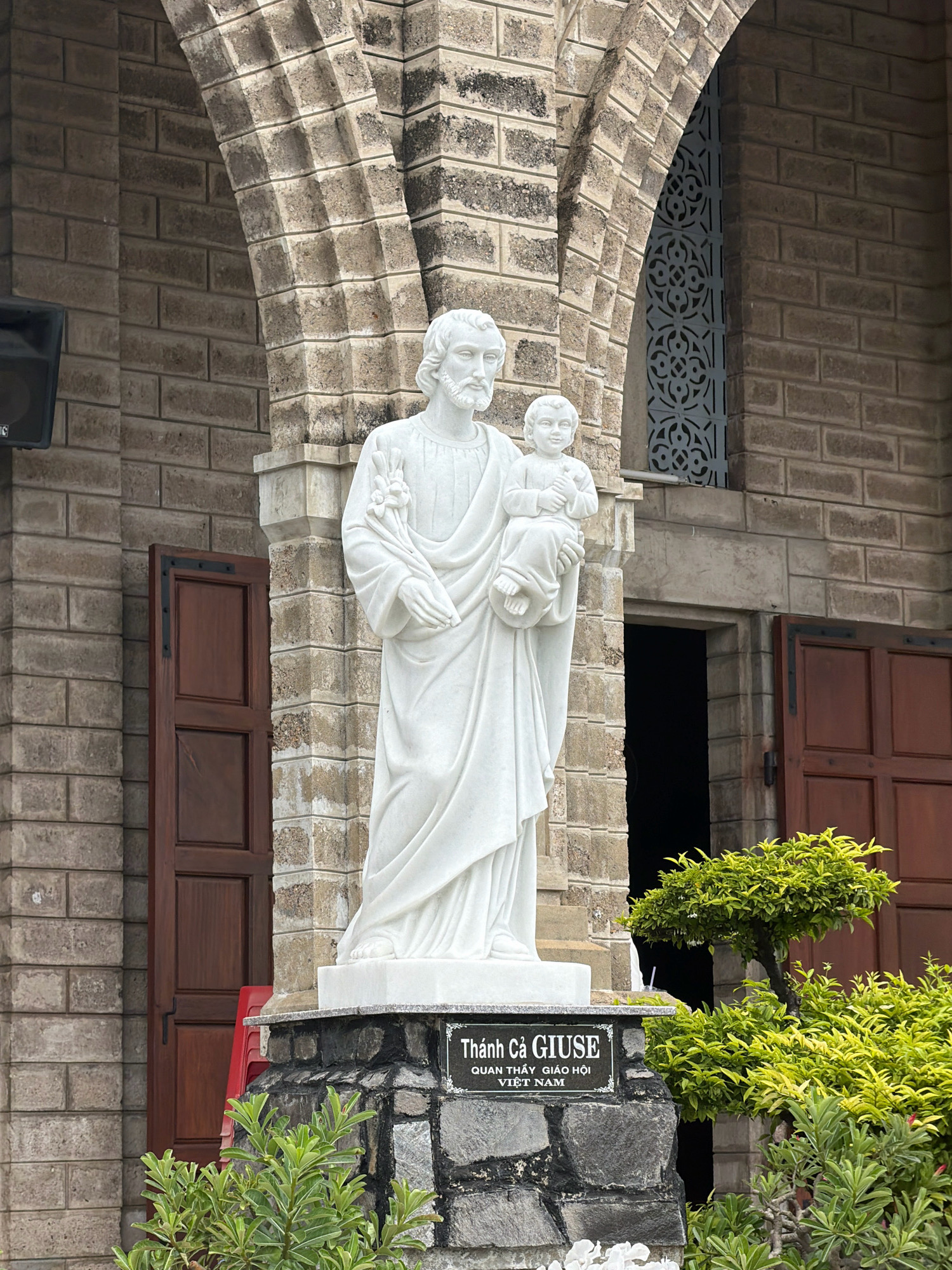 A white marble statue of Saint Joseph holding the child Jesus stands prominently in front of a stone-clad building in Nha Trang, Vietnam. The statue is positioned on a stone base with a black plaque bearing Vietnamese text. Behind the statue, the building features a large arched entrance with dark wooden doors. To the left of the statue, another dark wooden door is partially ajar, revealing a dark interior. Green foliage, including a small tree and shrubs, surrounds the base of the statue and the building. The architectural style of the building suggests it might be a church or religious institution. The lighting indicates an overcast or shaded day.