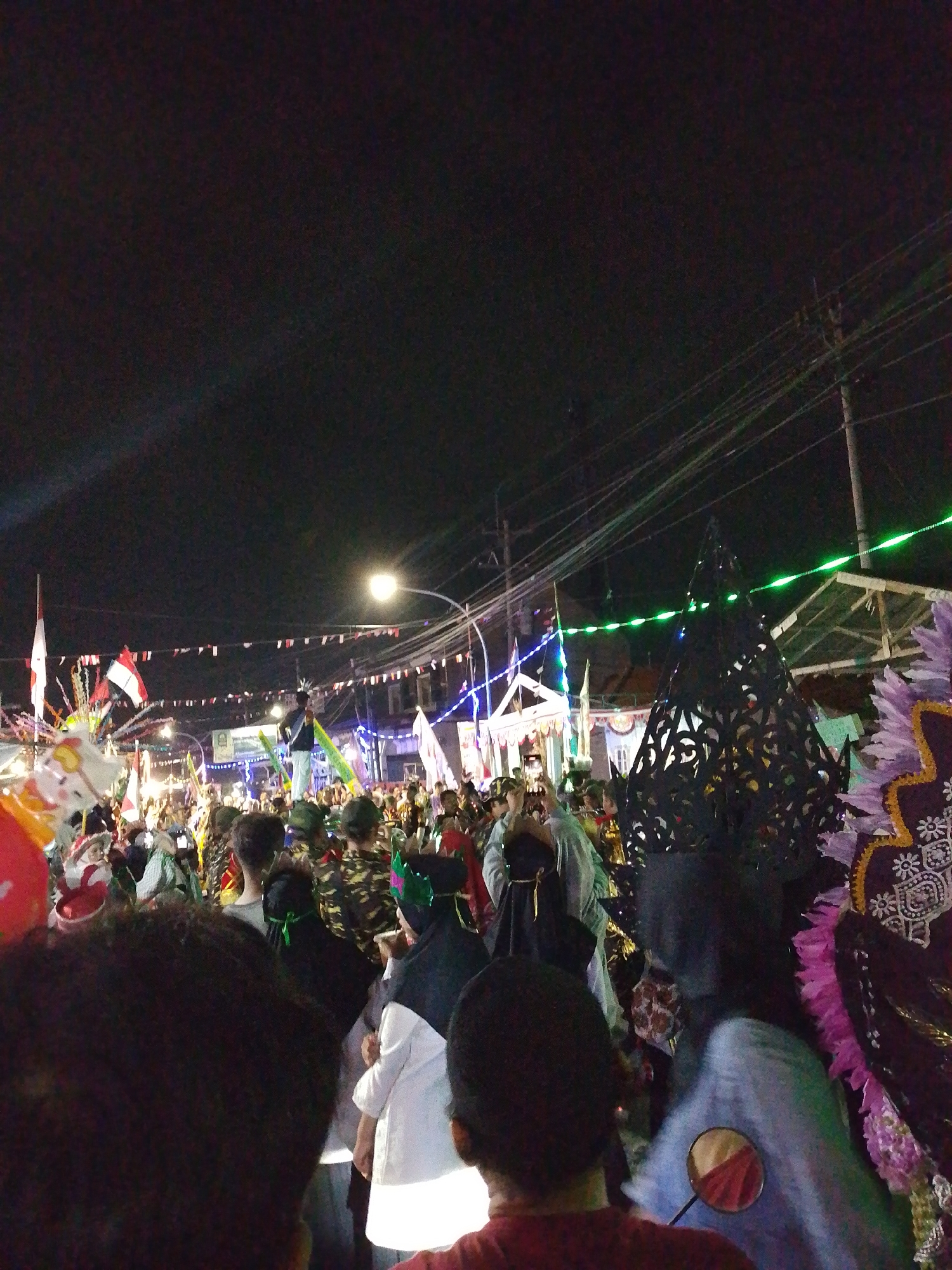 The image captures a nighttime street scene in Lawatan, Indonesia, during what appears to be a festive event. A large crowd of people are gathered, many of them looking towards a central area where a figure is standing on top of what seems to be a raised platform or vehicle. Numerous flags, including the Indonesian flag, are visible throughout the crowd and strung across the street. Decorative lights, some green and some white, illuminate the scene. Several people are holding up their phones, presumably to take pictures or record the event. In the foreground, the backs of several individuals are visible, suggesting the viewer is part of the crowd. The overall atmosphere is one of celebration and community engagement.