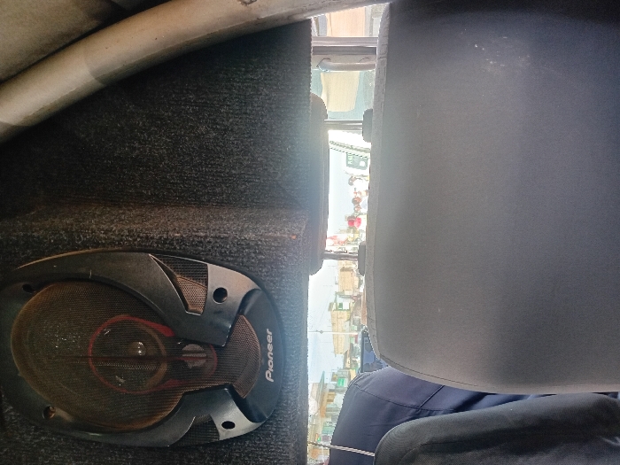 The image presents an interior view from a vehicle looking out onto a street scene in Naro Moru, Kenya. In the immediate foreground on the left, a dark grey, oval-shaped Pioneer brand car speaker, featuring a mesh grille and silver-colored structural accents, is mounted on a dark, textured, possibly carpeted panel. Dust is visible on the speaker and surrounding panel. The central midground is occupied by the dark grey fabric backrest and integrated headrest of a vehicle seat, viewed from behind. A light-colored, possibly metallic, structural pillar or bar extends vertically on the far right. Through the vehicle's rear window, a sunny urban environment is visible. Distant buildings, including multi-story structures with light-colored facades and various roofs, line a street. Multiple individuals are discernible in the distance, appearing to be walking or standing on the street. Vehicles are present, with one white truck partially visible bearing the text "PLANT" on its side. The sky above the scene appears bright and hazy.