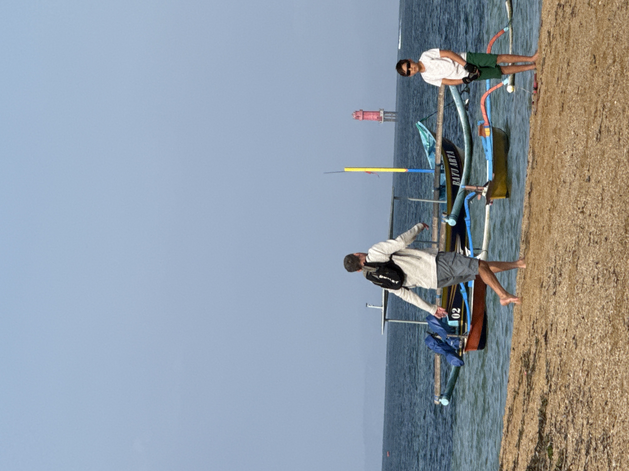 The image depicts a beach scene in Denpasar, Indonesia. A man with a backpack walks on the sandy shore towards the water, where several traditional outrigger boats are moored. A young boy stands on the beach near the water, looking towards the camera. In the distance, a red lighthouse is visible in the ocean. The sky is clear and blue, with calm water.