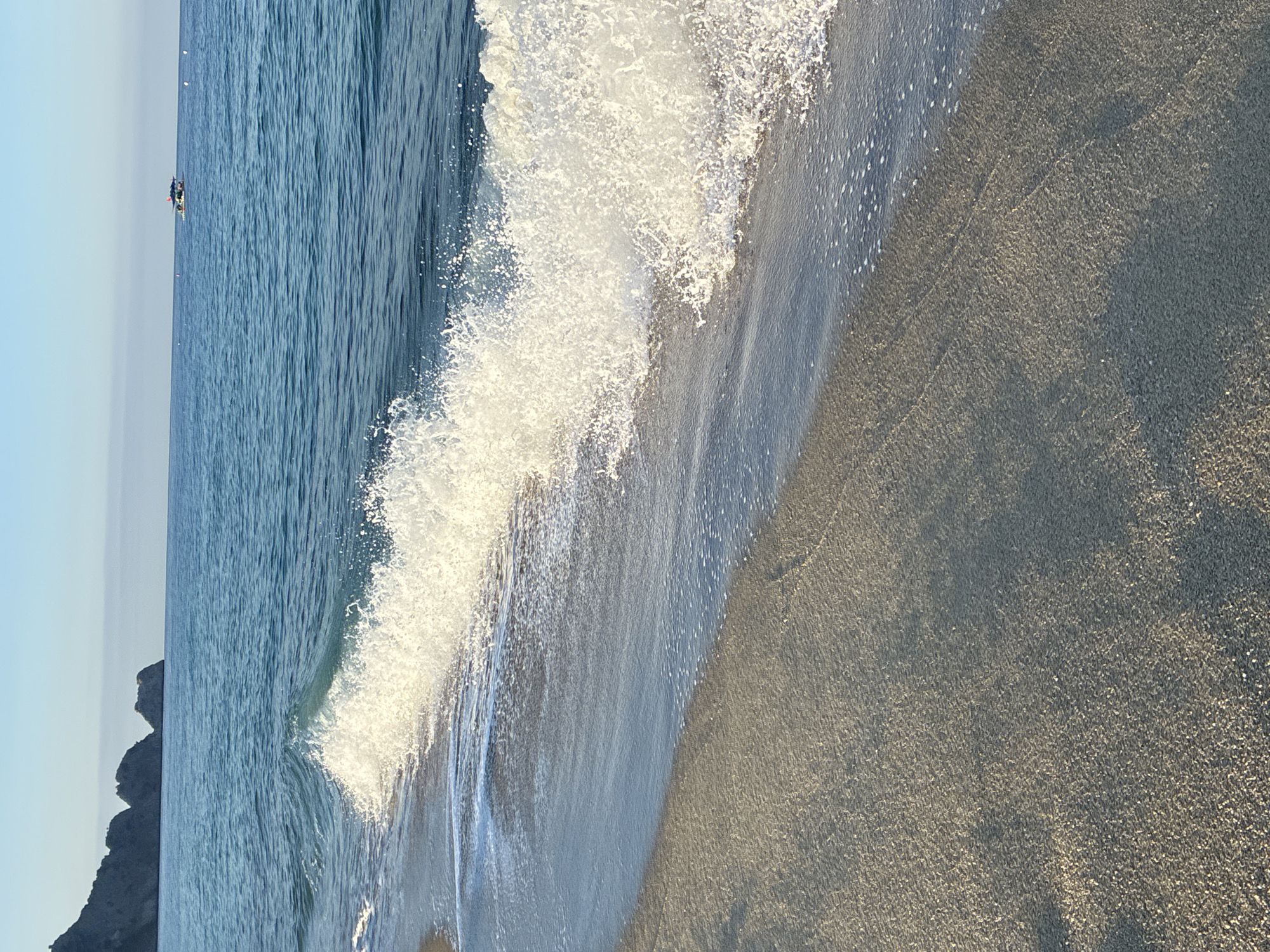 The image shows a view of the sea and beach. In the foreground, there is wet sand with shallow water lapping onto it. A wave is breaking, creating white foam and spray. The sea is a deep blue, and in the distance, there are rocky formations along the coastline. A small boat with a canopy is visible on the water. The sky is clear and blue. The scene depicts a tranquil beach environment, likely in Alanya, Turkey, given the rocky coastline which is characteristic of the region.
