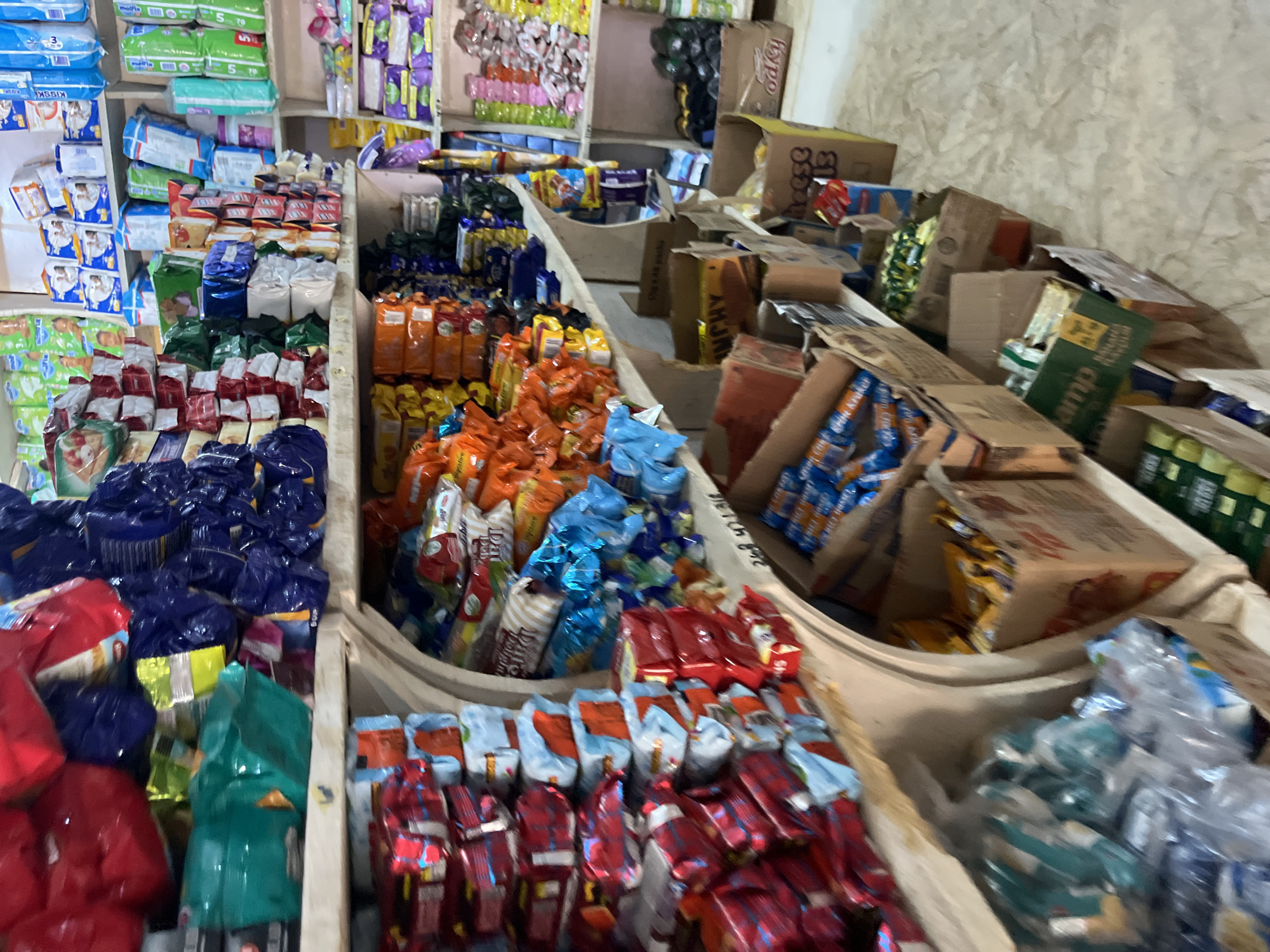 The image depicts an interior view of a small retail shop in Bukuru, Nigeria, showcasing shelves overflowing with various packaged consumer goods. The shop appears to be well-stocked, with products neatly arranged on wooden shelves and in cardboard boxes. The items are predominantly food products and household essentials, including snacks, biscuits, cereals, beverages, and personal care items. The overall impression is one of a busy marketplace with a wide variety of goods available to customers. The lighting is adequate, illuminating the products and shelves clearly. The environment suggests a typical Nigerian local market setting.