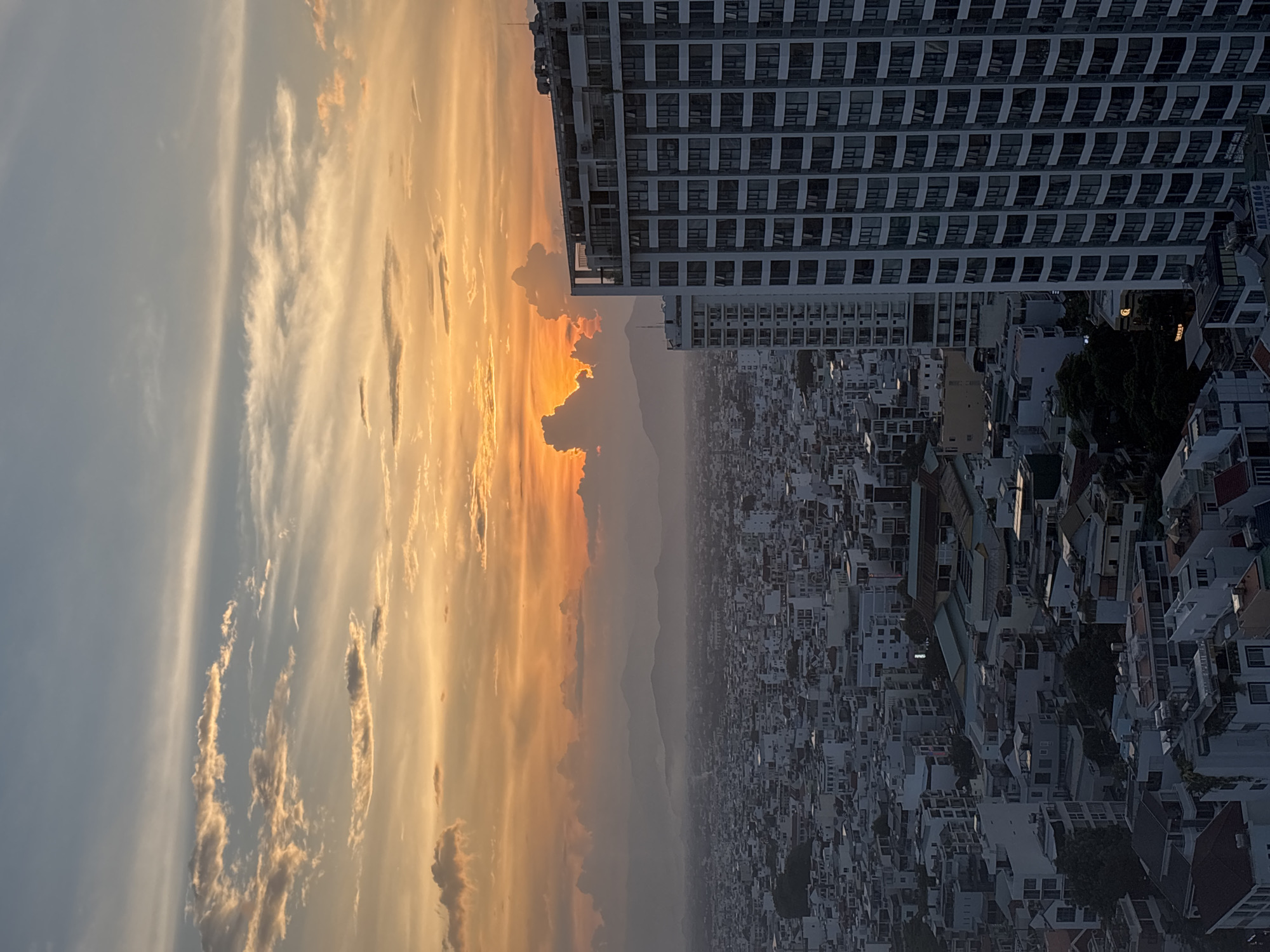 The image depicts a panoramic view of the city of Nha Trang, Vietnam, at sunset. The sky is a vibrant gradient of orange, yellow, and blue, with scattered clouds illuminated by the setting sun. In the foreground, a modern high-rise building dominates the right side of the frame, its numerous windows reflecting the warm hues of the sky. Beyond the building, the city sprawls out, a dense tapestry of smaller buildings with white facades and dark rooftops. In the distance, a range of hazy mountains forms a silhouetted backdrop against the colorful sky. The overall impression is one of urban development set against a dramatic natural landscape.