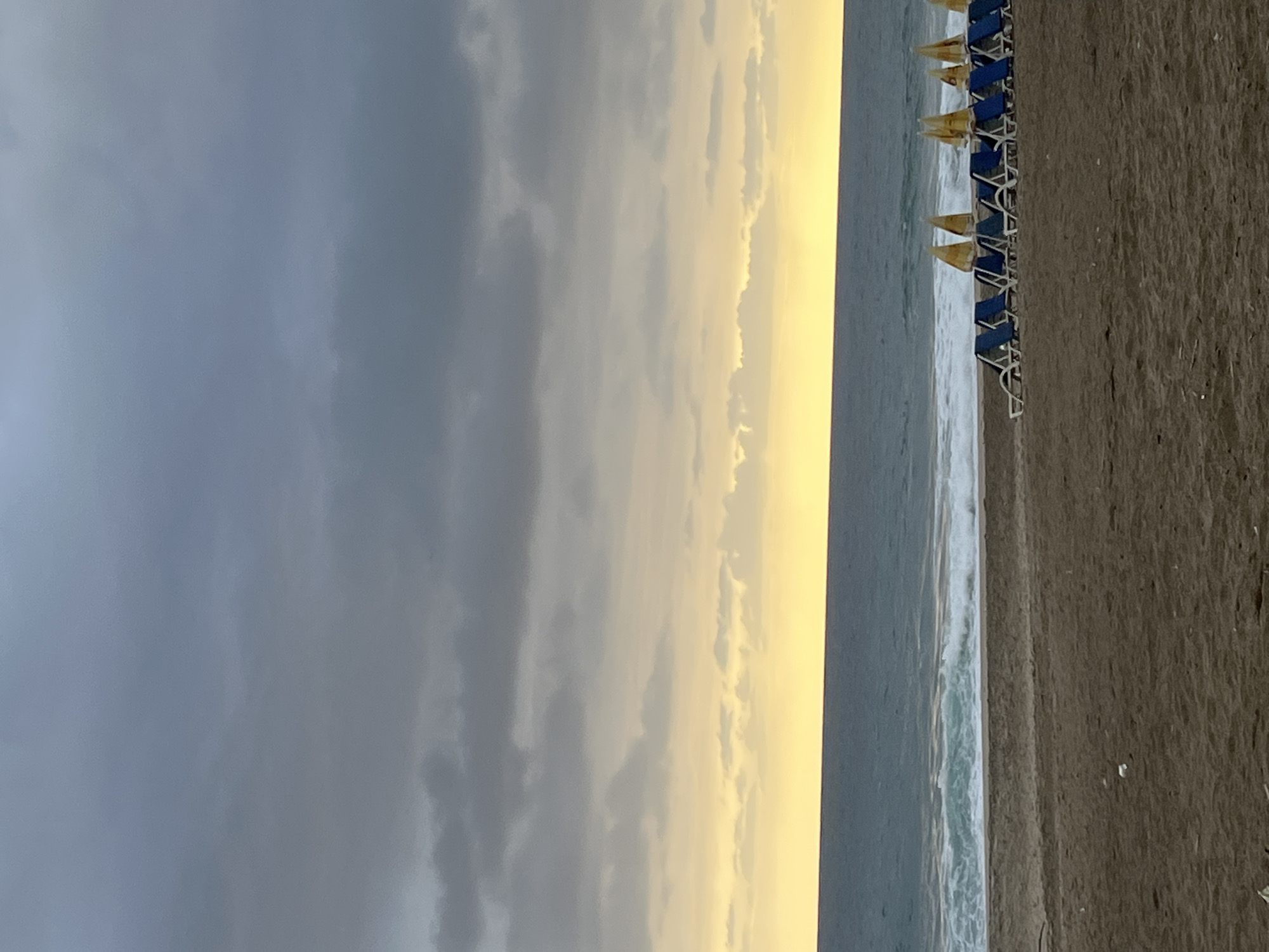 The image shows a beach scene in Alanya, Turkey, taken at what appears to be dusk or dawn. The sky is filled with dark grey clouds on the left, transitioning to lighter, yellowish clouds towards the center and right. The sea is visible as a narrow strip, with gentle waves lapping onto the shore. Along the shoreline, there are rows of neatly arranged beach chairs with blue frames and yellow canopies. The beach itself is composed of dark sand. No people are visible in the frame.
