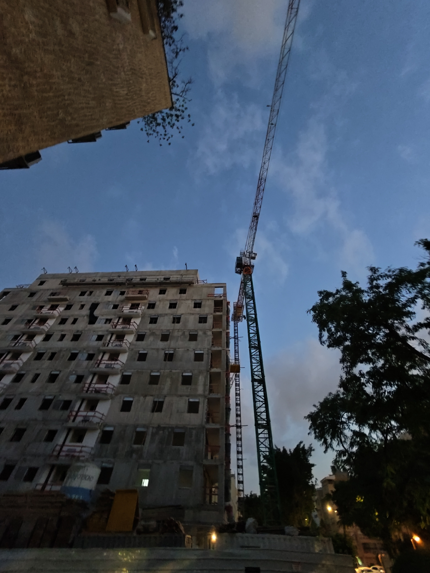 An outdoor, low-angle, wide shot captures a construction site in Petah Tikva, Israel. A towering green construction crane dominates the right side of the frame, its long arm extending diagonally upwards into a twilight sky filled with scattered clouds. To the left of the crane, a multi-story concrete building, also under construction, rises vertically. The building's facade features numerous dark, rectangular openings that appear to be windows or doorways, with some balconies visible. Red railings are present on some of these balconies. Below the main building, the scene transitions to a ground-level view of construction materials, including what appear to be stacks of wood and a yellow object. Dark green foliage from a nearby tree partially obscures the right side of the image. The overall lighting suggests it is either dawn or dusk.