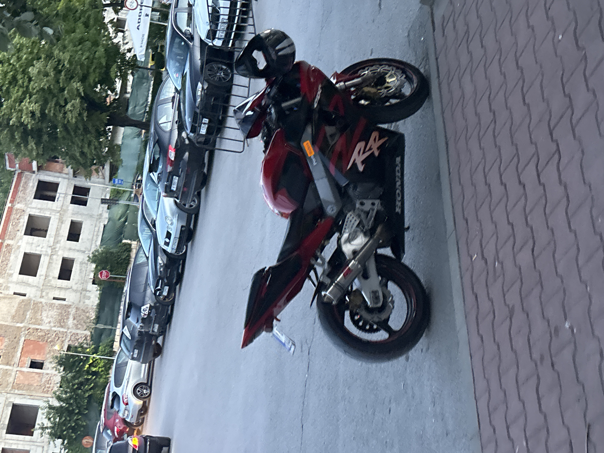 A red and black Honda motorcycle is parked on an asphalt surface in front of a sidewalk paved with reddish-brown interlocking bricks. The motorcycle is facing slightly to the right and is the primary subject of the image. A black helmet rests on the handlebars. In the background, several cars are parked in a row, including a black BMW and a white BMW. Beyond the cars, a multi-story building under construction with exposed brickwork and scaffolding is visible. Trees with green foliage are present behind the building, and a white tent with text is also in the background. A red stop sign is mounted on a pole to the left of the building. The scene appears to be an outdoor urban environment, possibly a parking area. The location is Shumen, Bulgaria.