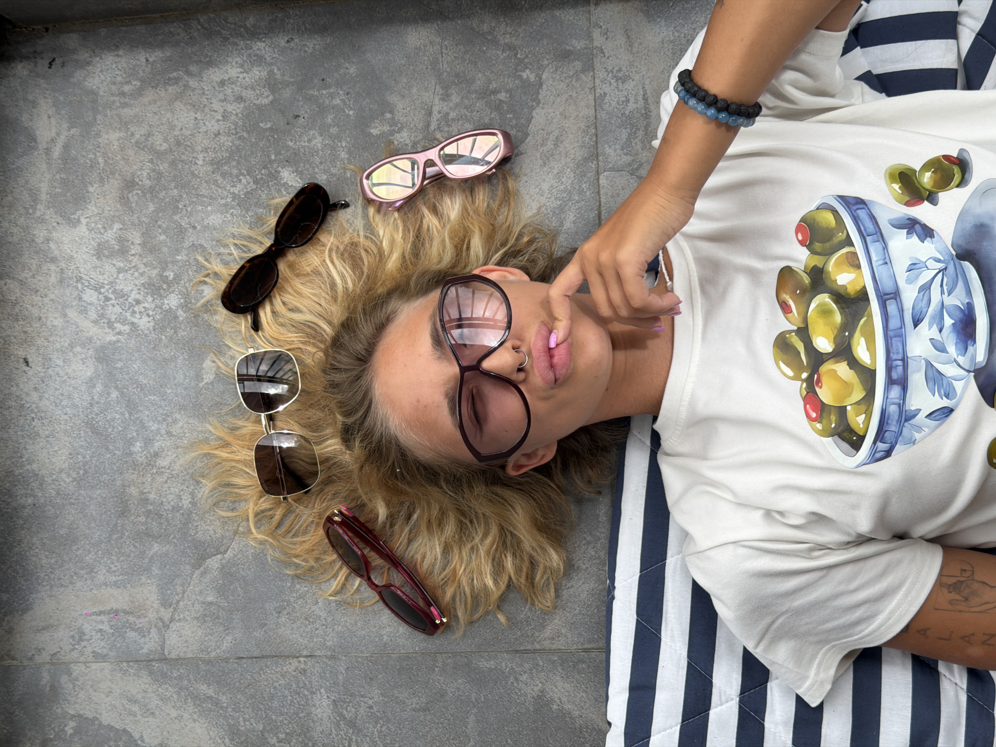 A young woman with blonde, wavy hair is lying on her back on a tiled surface, with her head and upper torso filling the frame. She is wearing a white t-shirt with a graphic print of olives in a bowl. Several pairs of sunglasses are scattered around her head: a pair of gold-rimmed aviators, dark tortoiseshell cat-eye glasses, burgundy framed sunglasses, and pink-framed futuristic sunglasses. The woman is looking directly at the camera with a playful expression, her lips pursed as if about to kiss. She is wearing a silver nose ring and has light purple nail polish on her fingers. A striped blanket is visible beneath her body, partially covering the tiled ground. The setting appears to be an outdoor urban environment, possibly a patio or courtyard in Tbilisi, Georgia. The lighting suggests it is daytime.
