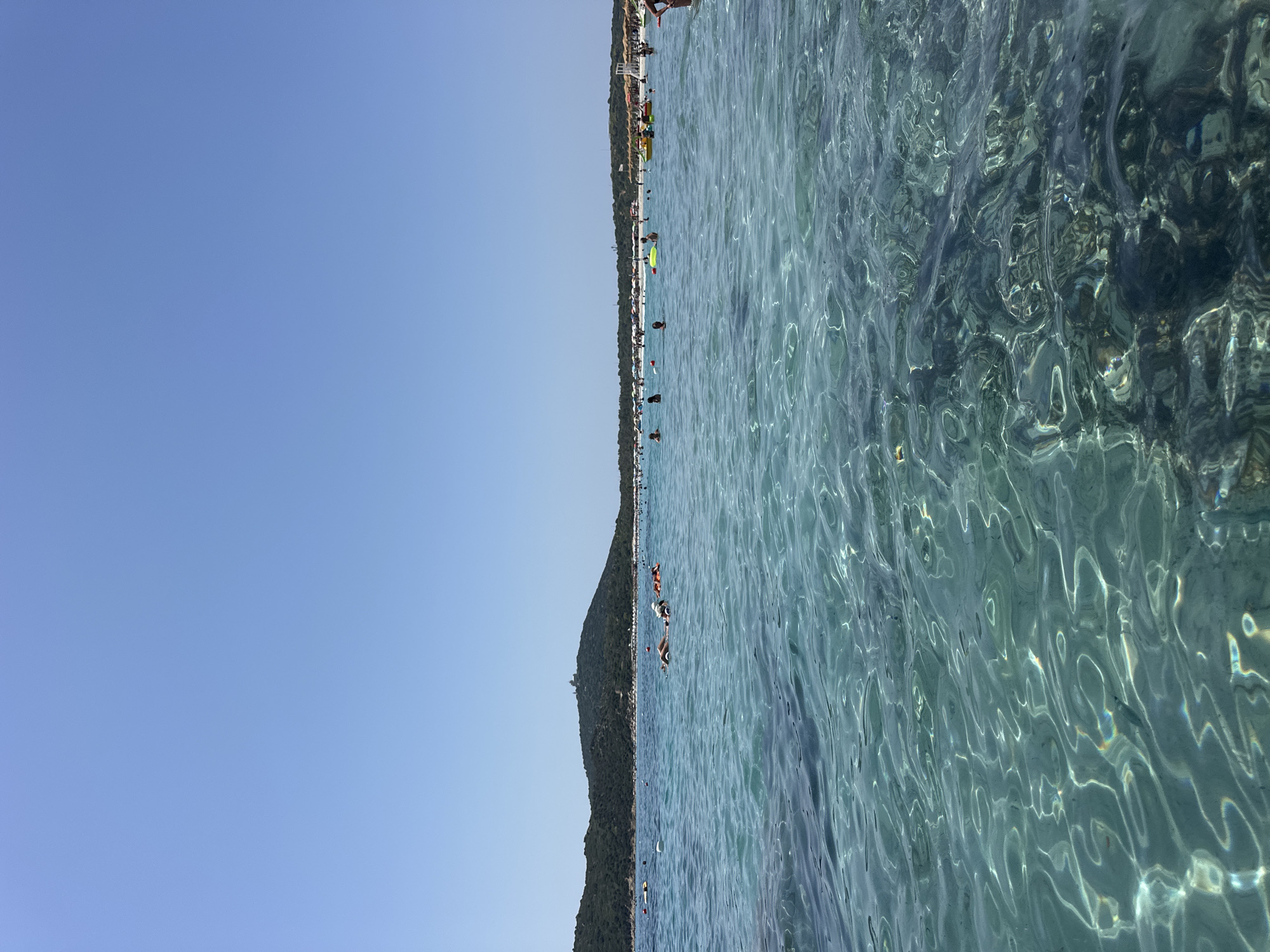This is a daytime, eye-level, vertical shot of the sea and a beach at Porto Giunco, Villasimius, Sardinia, Italy. The foreground is dominated by clear, turquoise water with ripples reflecting sunlight and revealing the sandy bottom and rocks beneath. In the midground, numerous people are in the water, some swimming, others on inflatable rafts, with buoys marking off areas. A distant coastline features a densely vegetated hill with a tower-like structure on its summit. Beyond the water, a crowded beach is visible with rows of umbrellas and sun loungers. The sky is a clear, pale blue.