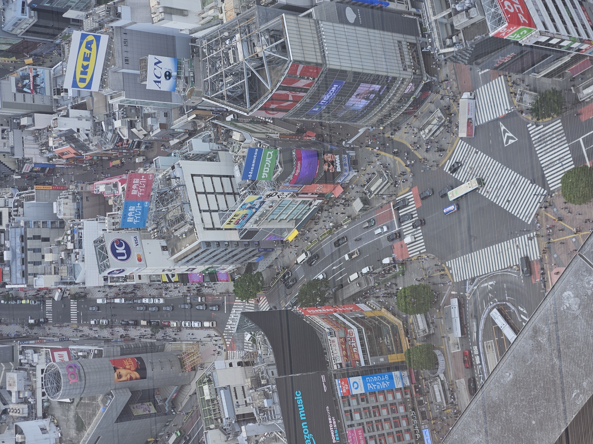 Here is a detailed description of the image:
The image is an aerial view of a bustling urban intersection, likely in a major Japanese city like Tokyo. The intersection is characterized by a dense crowd of pedestrians crossing in various directions, a phenomenon known as a scramble crossing. White stripes mark the pedestrian walkways on the dark asphalt of the road.
Buildings surround the crossing, many of which are covered with large, colorful advertisements and billboards featuring logos such as IKEA, ACN, UC VISA, Rakuten, DMM, ITSCOM, and Tsutaya. There are also advertisements for local Japanese products and services like salonpas. The architecture is modern, with a mix of glass-fronted structures and high-rise buildings.
Road traffic, including cars and buses, can be seen moving through the intersection, carefully navigating through the large crowd of pedestrians. The overall atmosphere is vibrant and energetic, reflecting the high density of activity in the area. A portion of the building, with steel structures and billboards, is visible on the right.