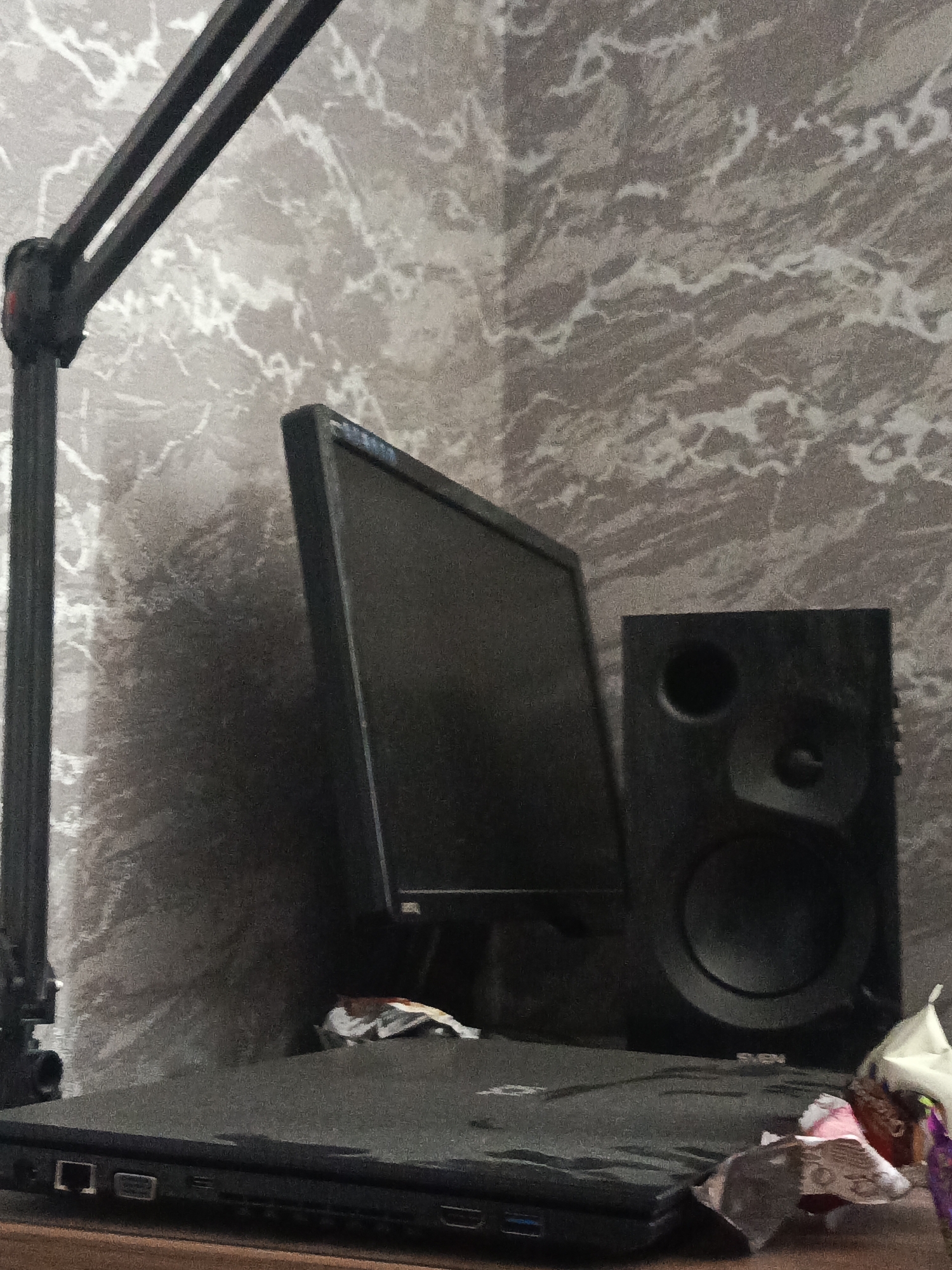 A desktop monitor, a speaker, and a laptop are set up on a surface against a patterned wall. The monitor is dark and rectangular, with a light blue strip along the top edge. To the right of the monitor is a black speaker with a circular woofer and a smaller circular tweeter. In front of the monitor and speaker is a closed black laptop. Some crumpled papers and wrappers are scattered on the laptop and the surface. A black desk lamp arm extends from the left side of the frame. The scene is dimly lit, with the wallpaper having a gray and white marbled pattern. The location is identified as Lesosibirsk, Russia.