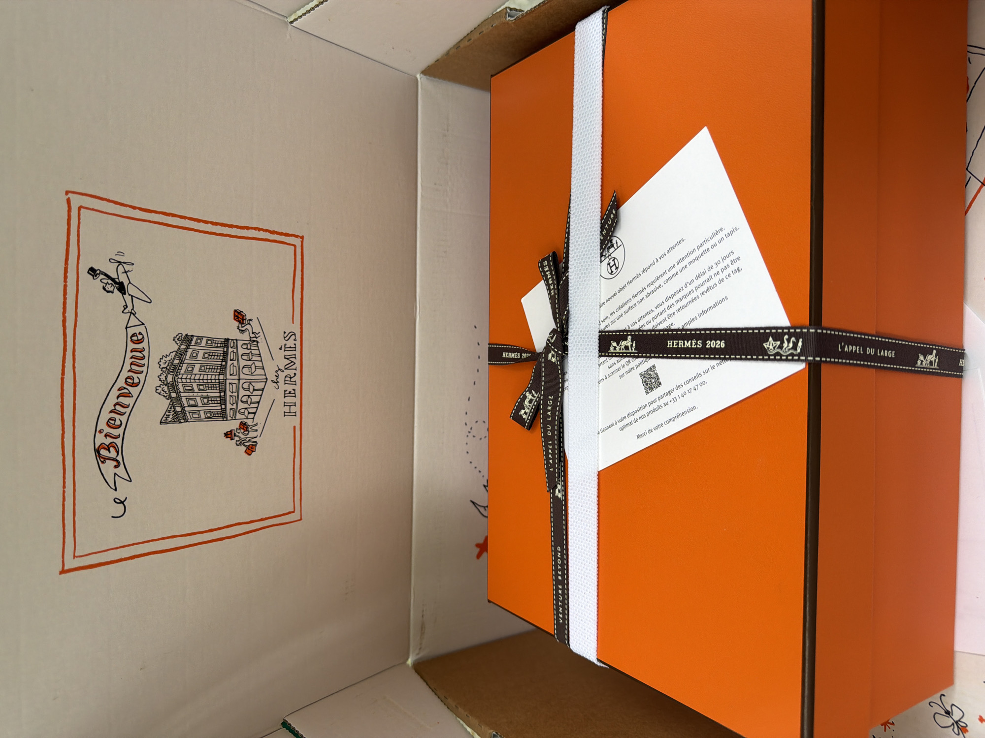 The image shows an open cardboard box with a white interior. Inside the box, an orange Hermès gift box is visible, tied with a dark ribbon featuring the Hermès logo and "L'APPEL DU LARGE" printed on it. The lid of the box is decorated with a hand-drawn illustration of a building and a banner that reads "Bienvenue chez Hermès." The scene is indoors, likely a home or office setting. There are no people or specific landmarks indicating Penzano, Italy.