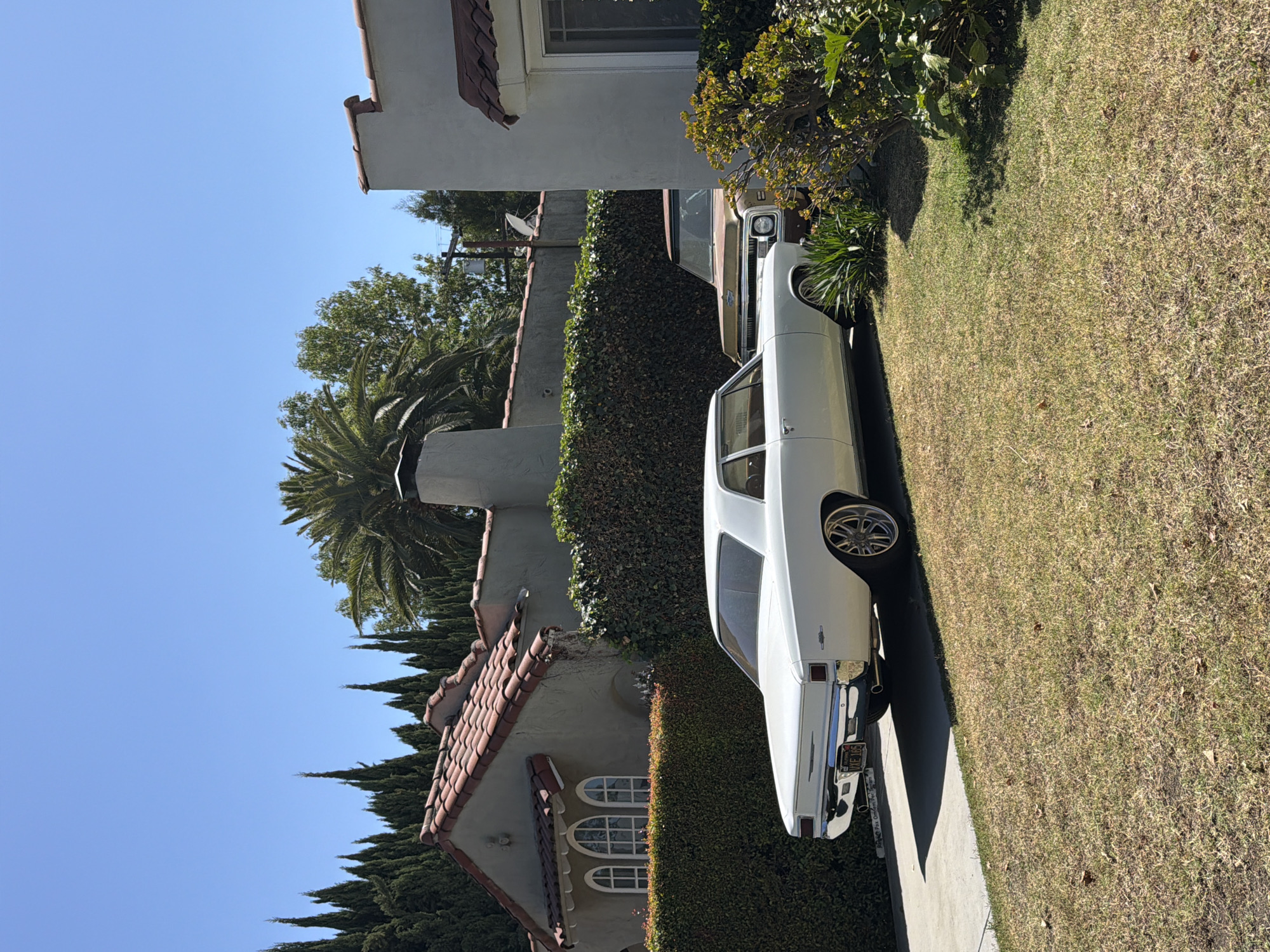 The image shows a white sedan parked on a paved driveway alongside a lawn in front of a residential property. The property has a white stucco exterior with a red-tiled roof, arched windows, and a tall hedge. A palm tree is visible in the background. The license plate on the sedan is partially obscured, but it is possible to read "AKE 06_" and "California". Based on architectural styles, foliage and license plate this is likely an urban residential neighborhood in Los Angeles, California. Another vehicle, likely a truck, is partially visible behind the parked sedan. The sky is clear and blue, suggesting a sunny day.