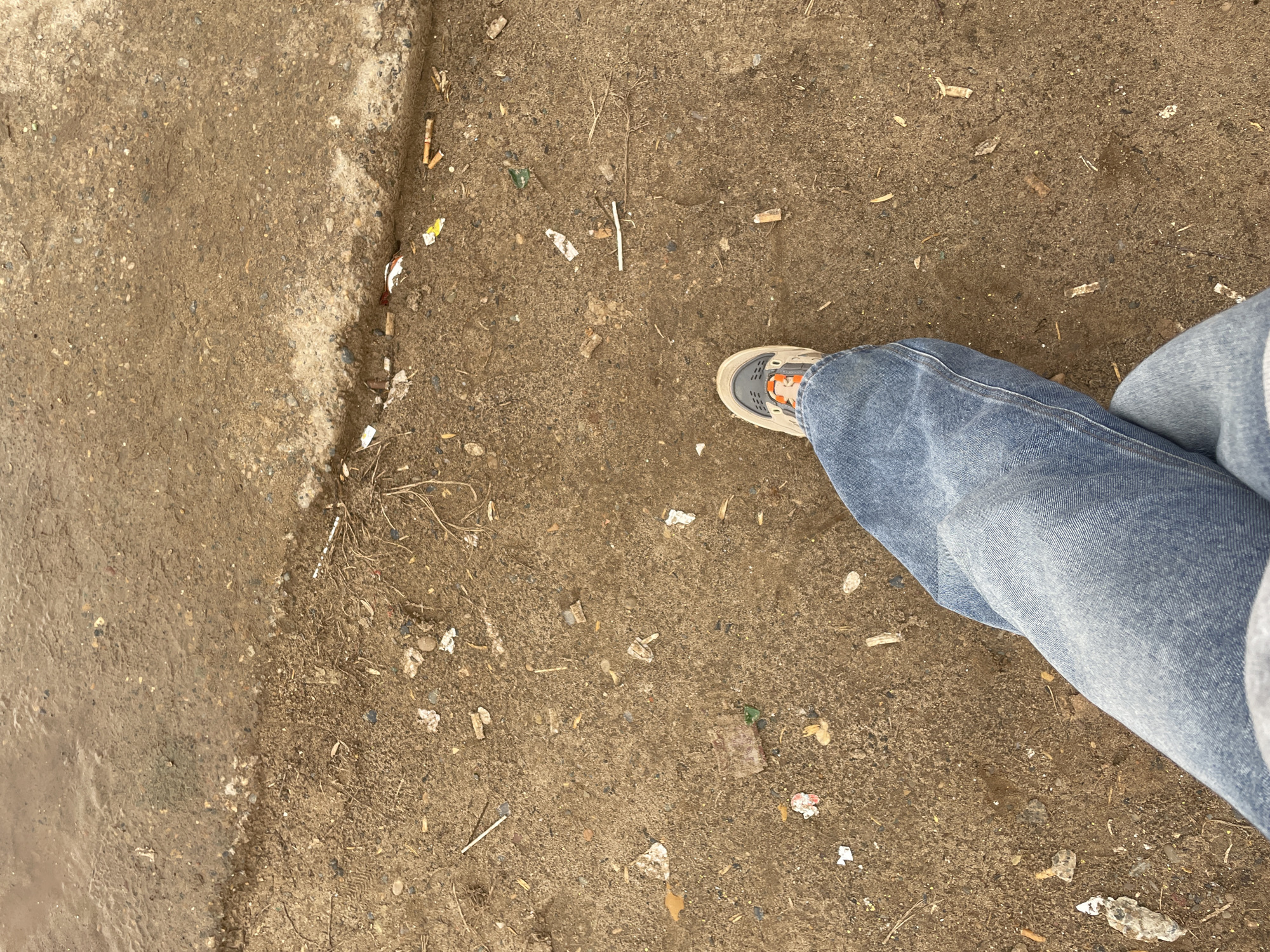 A ground-level shot shows a person standing on a dirty sidewalk in what appears to be a city environment. The sidewalk is composed of brown earth and gravel, with litter scattered across the surface, including cigarette butts, small scraps of paper, and other debris. Part of the sidewalk is wet and discolored. The person in the image is wearing light-wash denim jeans and a beige and orange athletic shoe. It is unknown if the image was taken in Ольховка, Russia.