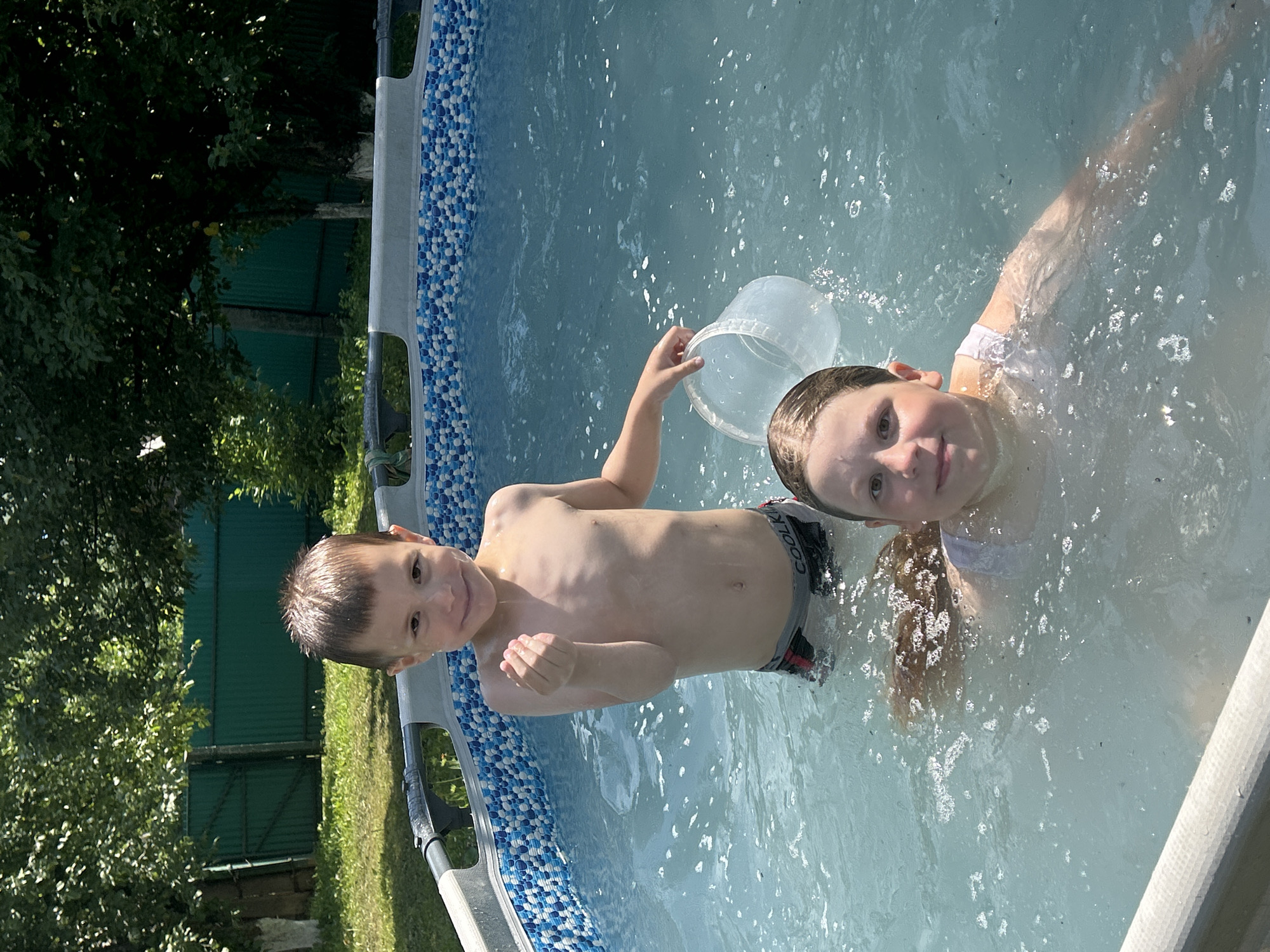 In this overhead shot taken on a sunny day in Фоки, Russia, two children are playing in a backyard swimming pool. The water is clear blue, with visible ripples and bubbles. The pool has a blue and white patterned liner.
On the left, a young boy with short brown hair and a bright smile looks directly at the camera. He is wearing dark swim trunks. His body is mostly submerged in the water, with his arms visible.
To the right of the boy, a younger girl with blonde hair is partially visible. She is wearing a white top. Her arm is raised, holding a clear plastic bucket. Both children appear happy and engaged in their play.
The background shows lush green trees and a dark green fence, suggesting the pool is located in a garden or yard. The bright sunlight indicates it is likely midday or early afternoon.