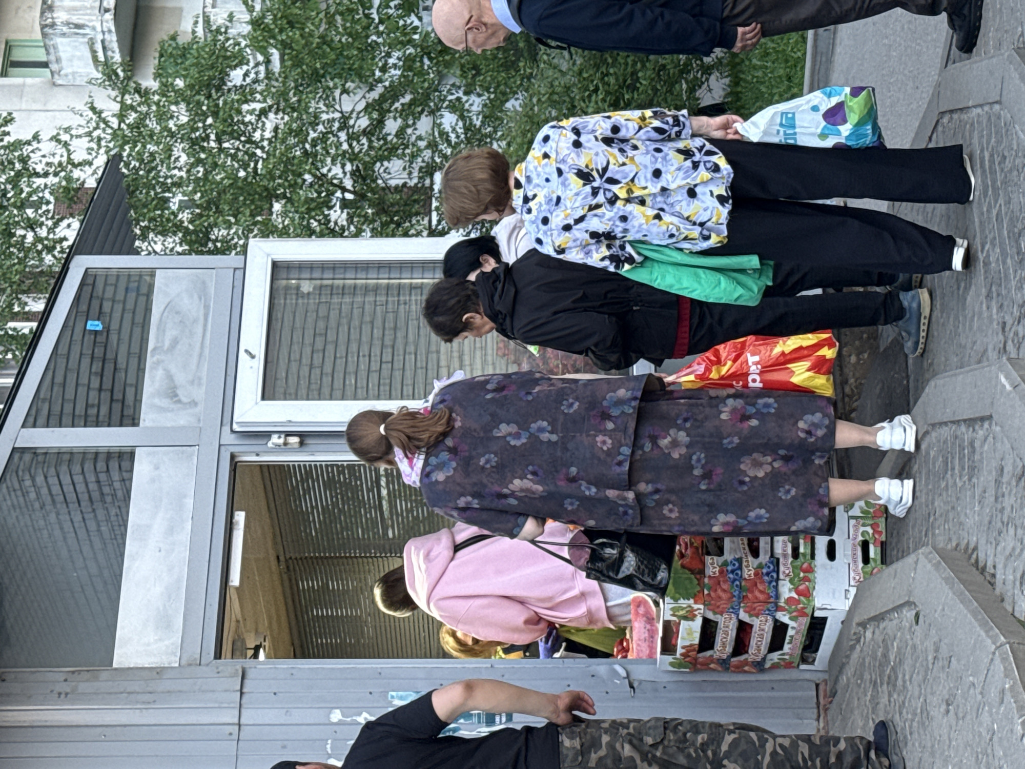 A scene outside a small commercial building in Moscow, Russia, features a line of people standing at a small fruit stand. An elderly man with a bald head and wearing a dark jacket is at the front of the line. A woman in a white shirt with black and yellow floral patterns and black pants stands behind him. Another woman in a black jacket follows. Then, a woman in a purple floral robe. Behind her is a woman wearing a pink hoodie. The fruit stand displays produce in cardboard boxes. One of the boxes is labeled in Russian, "Kubanskaya Yagoda." The building is grey and silver, with several windows. A tree is visible behind the building. The street is a mixture of pavement and concrete.