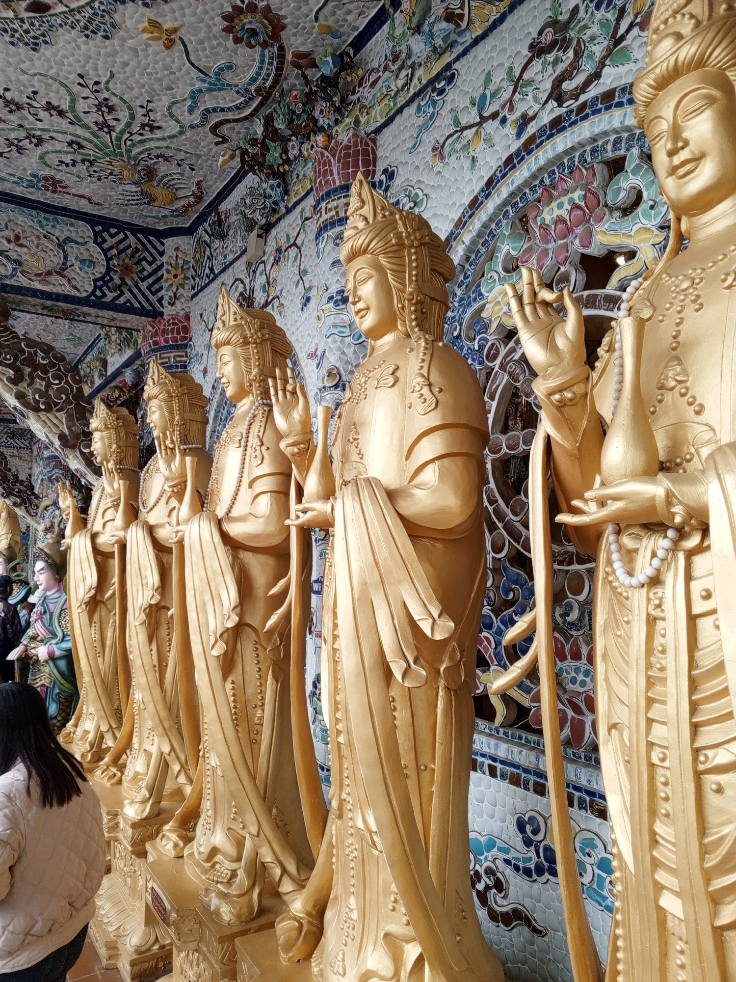 The image depicts a row of golden statues, likely of Buddhist deities, aligned along a wall adorned with intricate colorful mosaic tiles. The statues are standing upright, with their hands raised in various gestures. The wall behind them features a vibrant pattern of floral motifs and stylized clouds. In the foreground, the back of a person wearing a light-colored jacket is visible, suggesting a visitor to the location. The scene appears to be an interior space, possibly a temple or a religious site, given the statues and the ornate decoration. The location is identified as Xuân Thành, Vietnam, which is a coastal town known for its beaches and increasingly, its religious tourism.