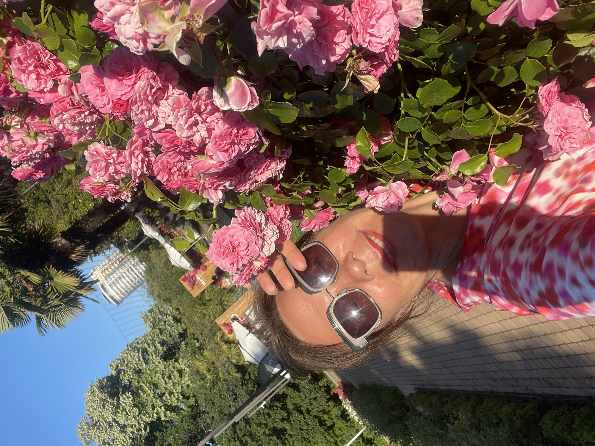 A woman takes a selfie in front of a bush of pink roses. She is wearing sunglasses and a pink and white tie-dye shirt. The background shows a pathway, lush green trees, and a modern building. This location is on the coast of the city of Sochi, Russia, on a sunny day.