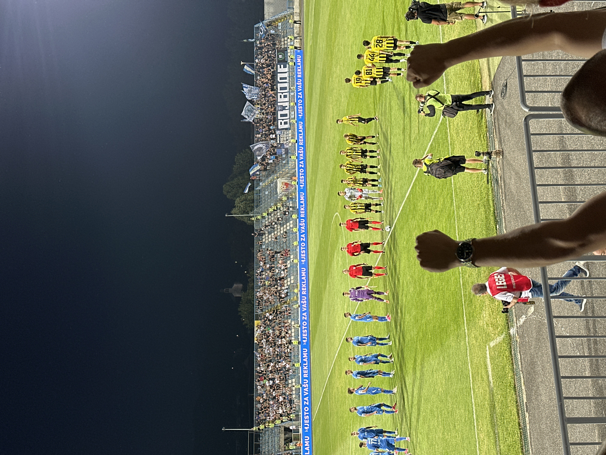 The image depicts a nighttime outdoor scene at a football stadium, likely in Nikšić, Montenegro, during a match. Two teams are lined up on the green pitch. One team, wearing blue uniforms, stands on the left side of the frame, while the opposing team, in yellow and black striped uniforms, is positioned on the right. Referees in red uniforms are also present on the field, one at the center of the yellow-striped team and another towards the right of the frame. Spectators fill the bleachers in the background, some holding flags. A banner with text, potentially advertising, runs along the front of the stadium seating. In the foreground, several individuals, including a photographer with a camera and a person with a camera on their shoulder, are visible. The overall atmosphere appears to be one of sporting event engagement.