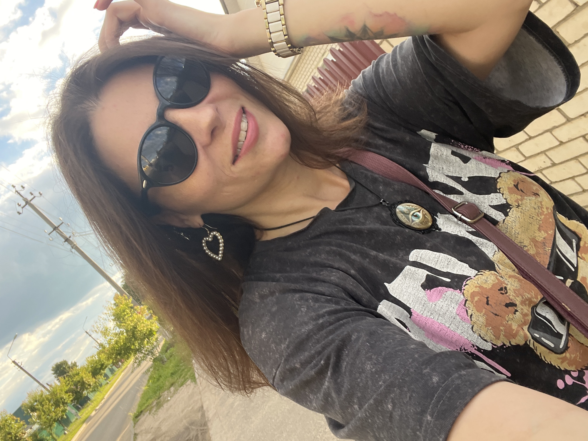 In Krasnoe-na-Volge, Russia, a woman with long brown hair, wearing black sunglasses, a dark grey graphic t-shirt, and a maroon strap across her chest, stands on a paved walkway next to a road. She is wearing heart-shaped earrings and a pendant necklace. A brick wall is visible behind her.