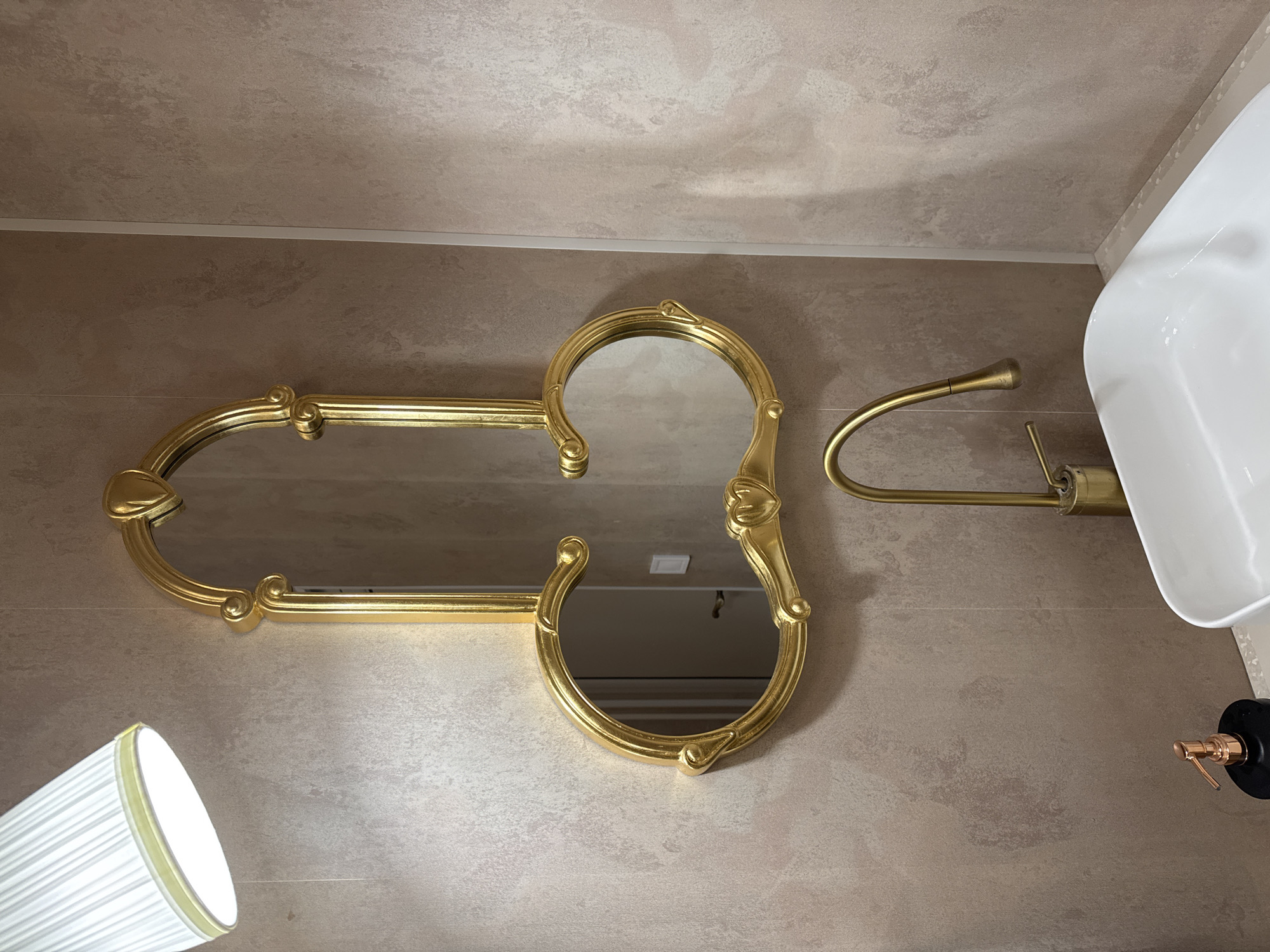 The image depicts a bathroom interior. The main focal point is a gold-framed mirror in an unusual shape, resembling two interlocked circles with extended horizontal elements. A gold towel rack is adjacent to the mirror. To the right, a white sink and soap dispenser are partially visible. At the bottom left corner is a lamp with a white shade. The wall behind the fixtures is covered in a beige textured material. The city and location are unknown.