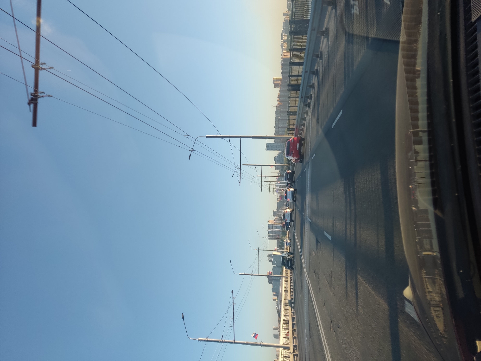 The image depicts a view from a vehicle driving on a multi-lane bridge, likely in Novosibirsk, Russia, given the presence of the Russian flag on a pole. The sky is clear and blue. Overhead, multiple power lines crisscross the scene, supported by tall poles. Several cars are driving on the bridge in the same direction. In the distance, a cityscape with numerous buildings is visible. The bridge has a metal railing on the right side, separating the road from the lower level. The road surface shows some wear and tear, with visible potholes. The sun appears to be low in the sky, casting long shadows across the road.